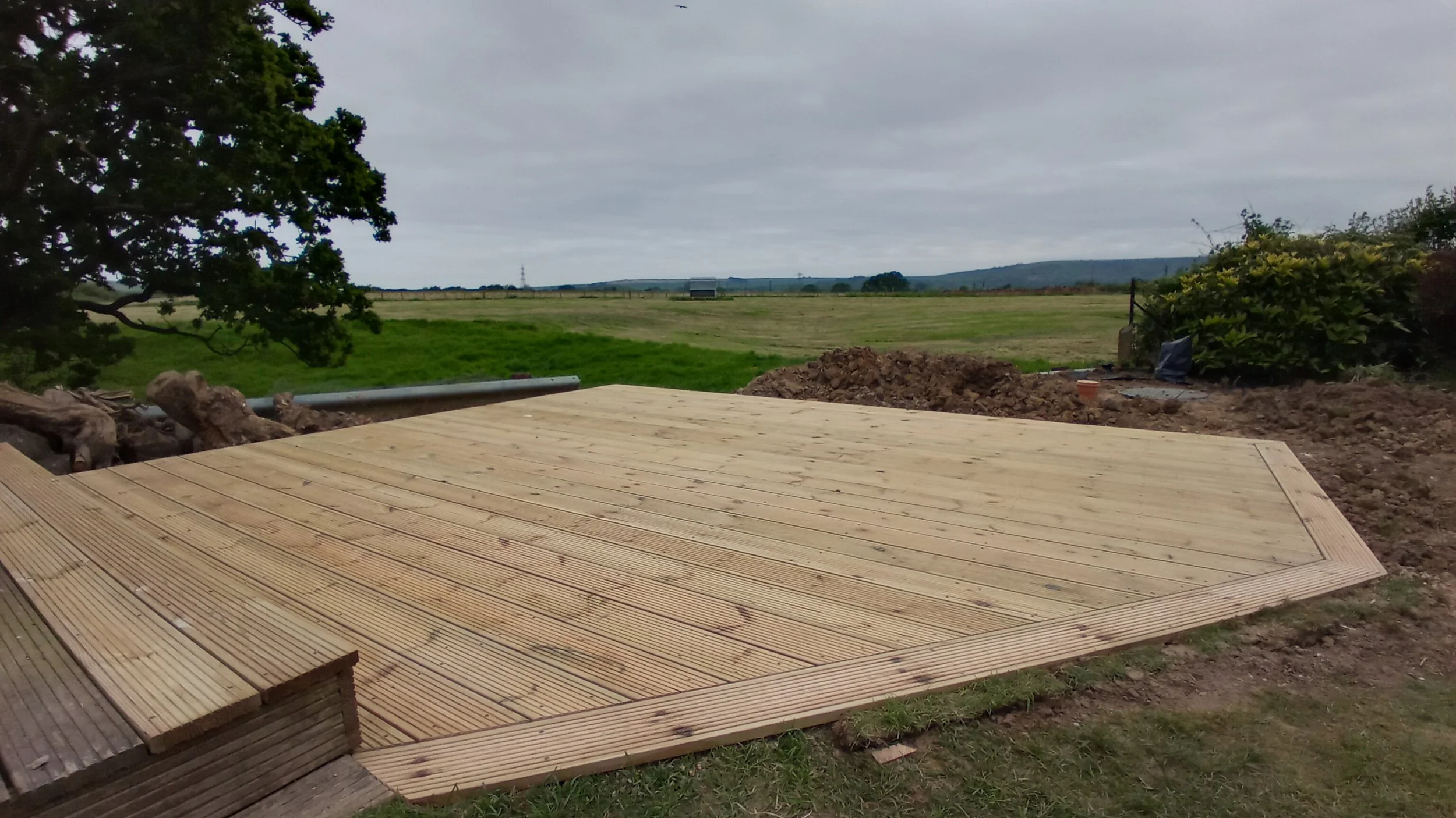 Newly constructed wooden deck in a rural area with green fields and trees under a cloudy sky.