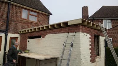 Construction site with a partially built brick wall extension and wooden beams overhead, ladders leaning against the structure, and a person working nearby.