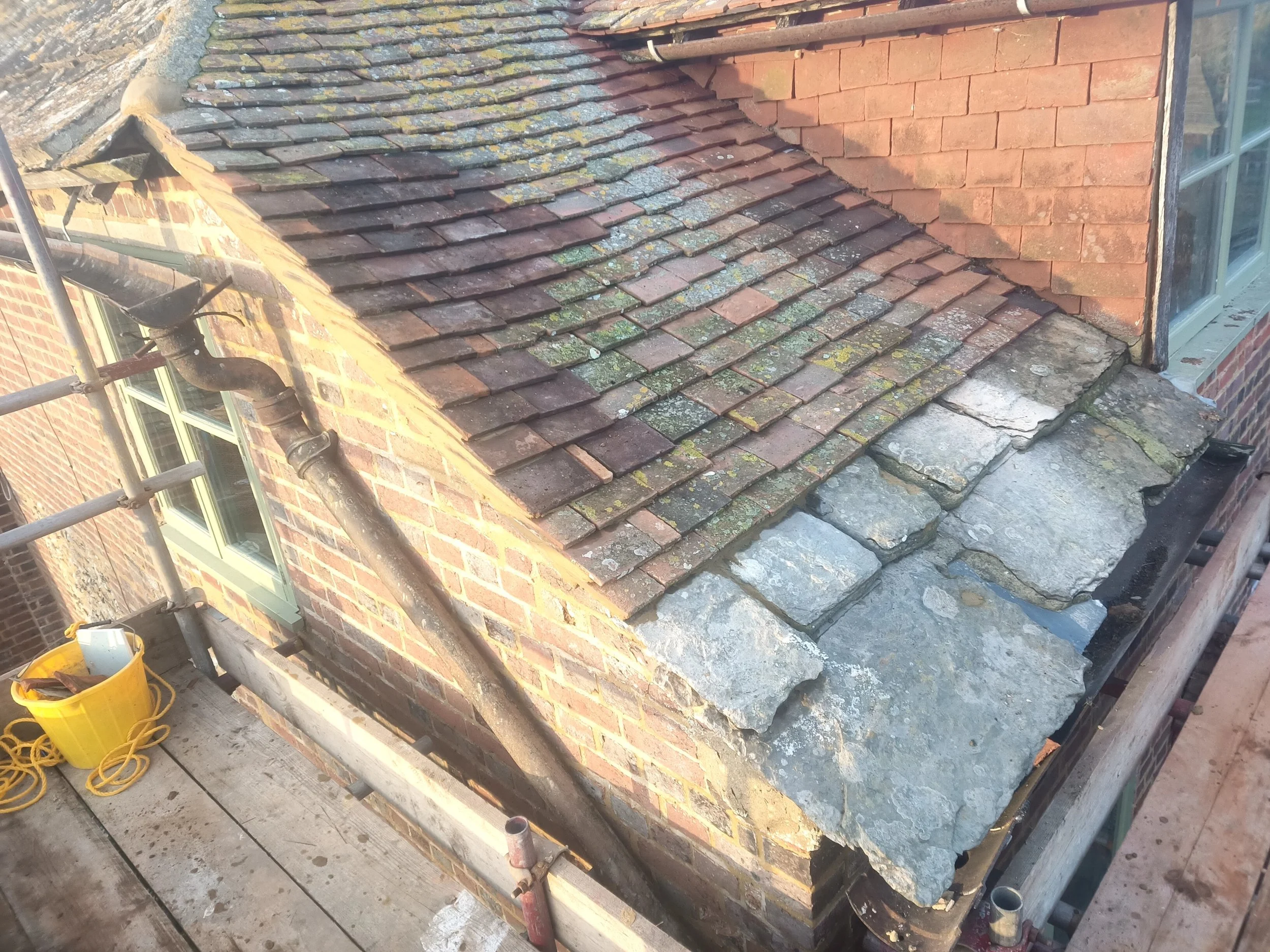 A roof with a mix of old and new tiles, some moss or lichen growth, and a section of damaged slate shingles. There is scaffolding and a yellow bucket on the surrounding platform.