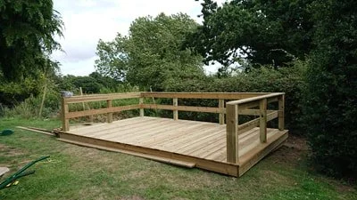 A newly built wooden deck with railing in a grassy yard surrounded by trees.