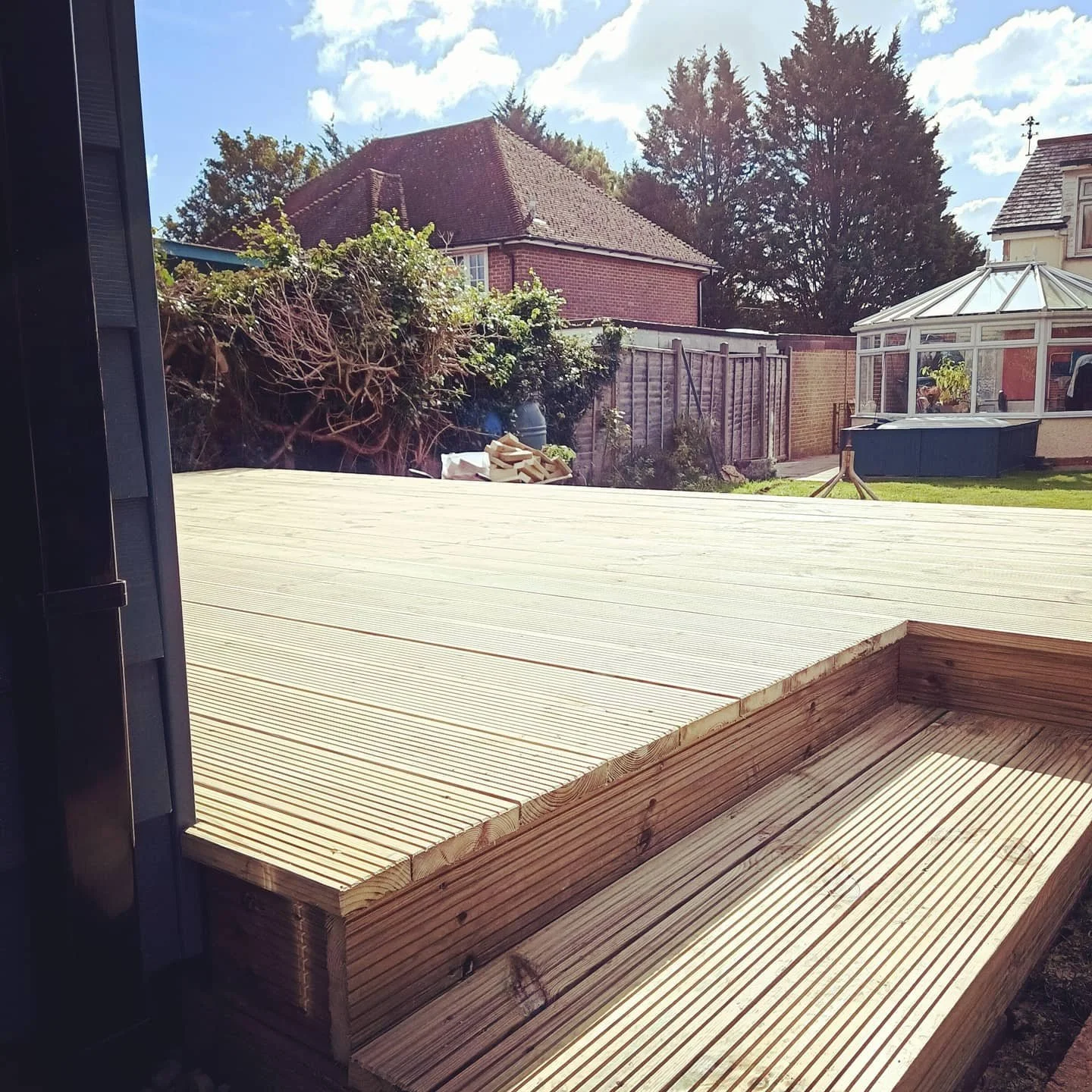 View of a backyard with a newly built wooden deck, a greenhouse, and houses with trees in the background.
