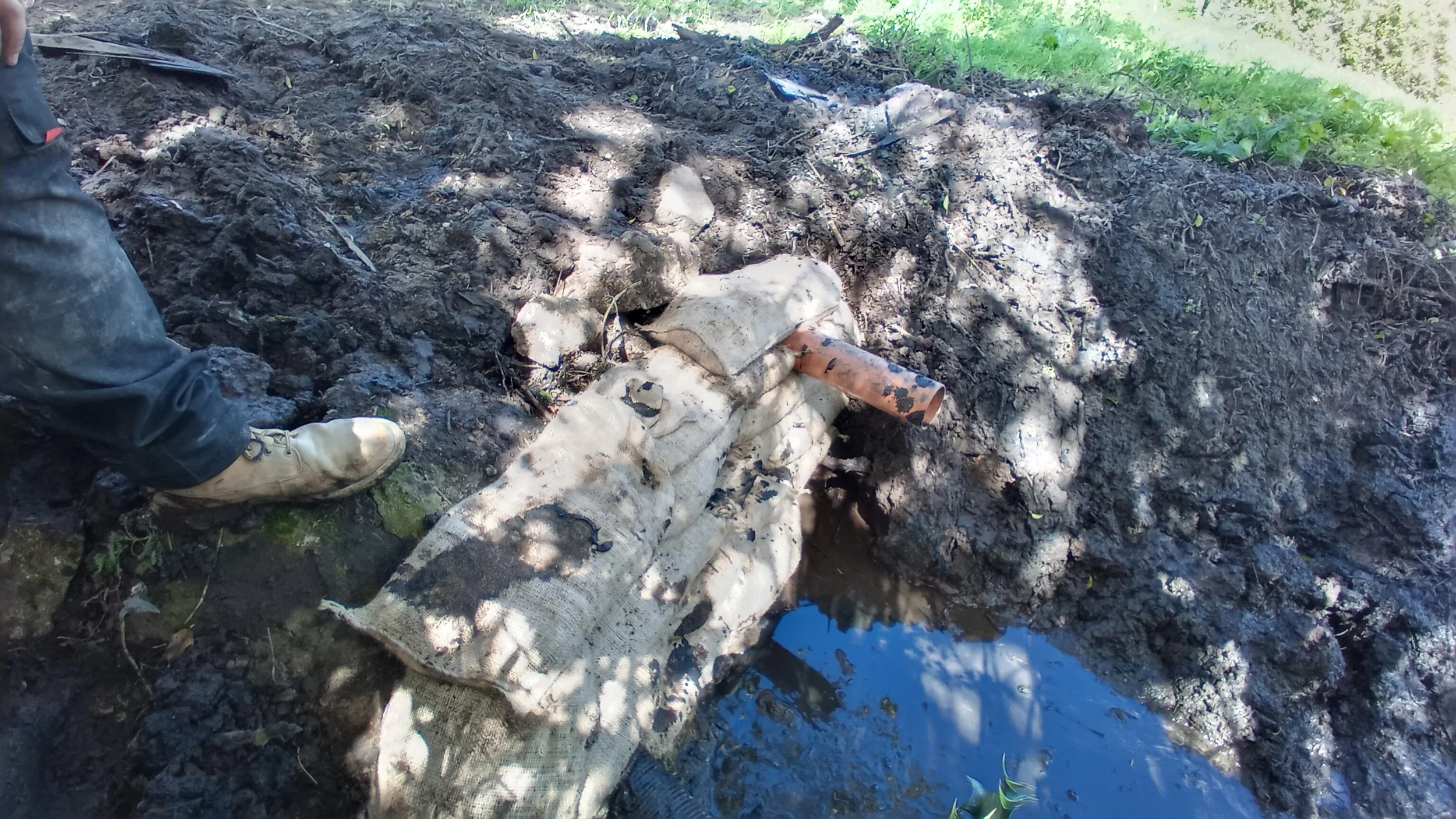 A blackened, burned area of ground with a partially buried, damaged pipe and a rusty pipe sticking out of a white, soot-stained bag or cloth, next to a small pool of water. A person's foot is visible on the left side, wearing a beige boot and black p