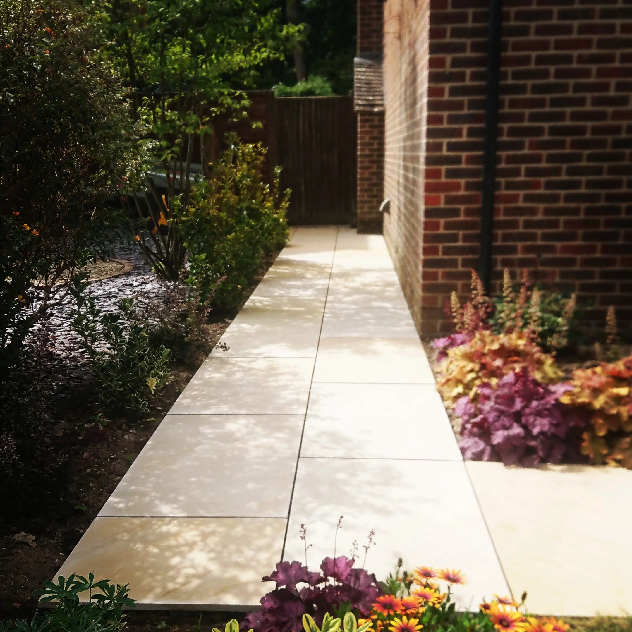 A Limestone Slab garden path next to a brick house, with vibrant flowers and green bushes on either side, and dappled sunlight through trees.