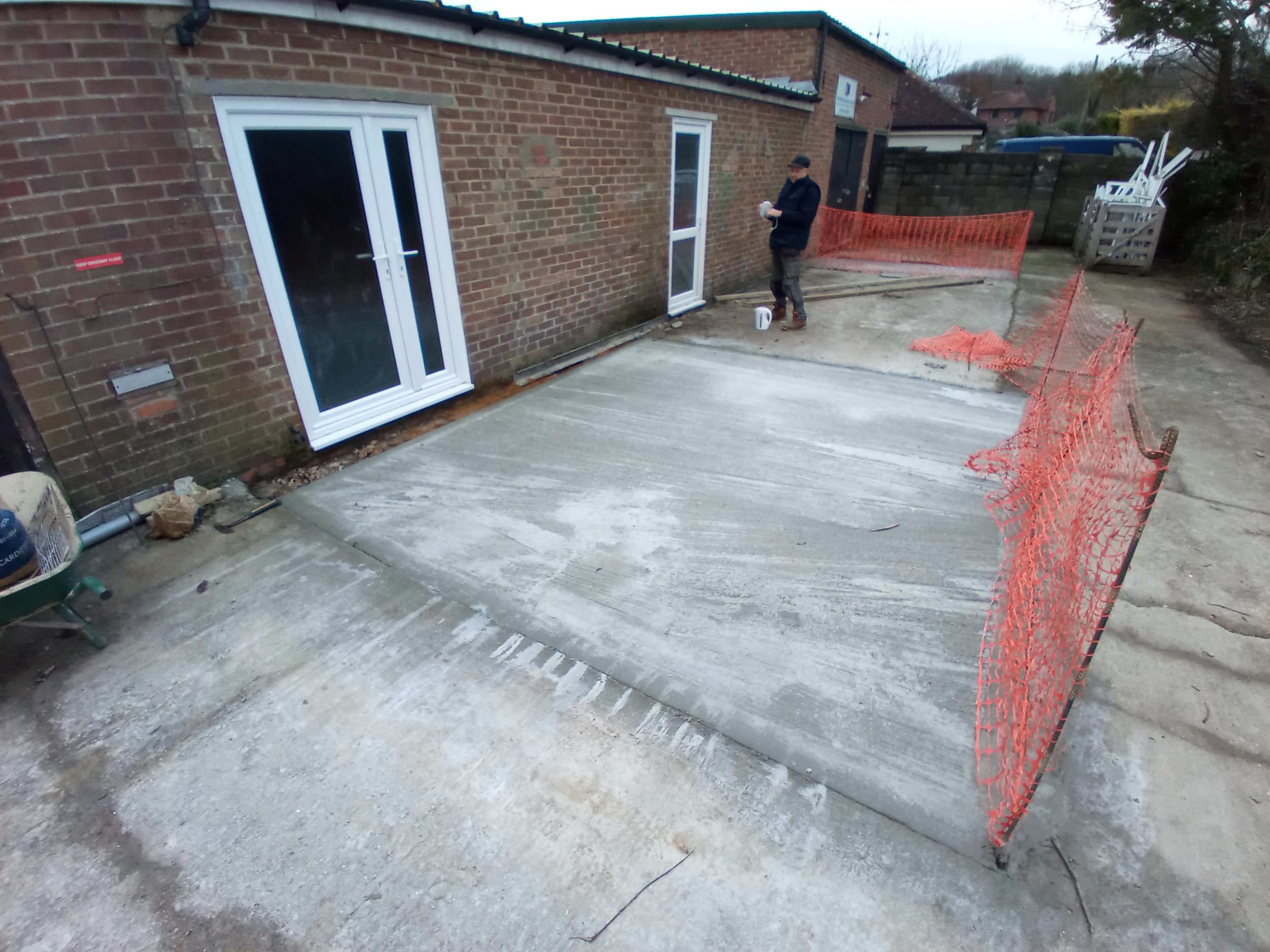 Construction site with a freshly poured concrete patio, orange safety fencing, and a man working near a brick building with white doors.