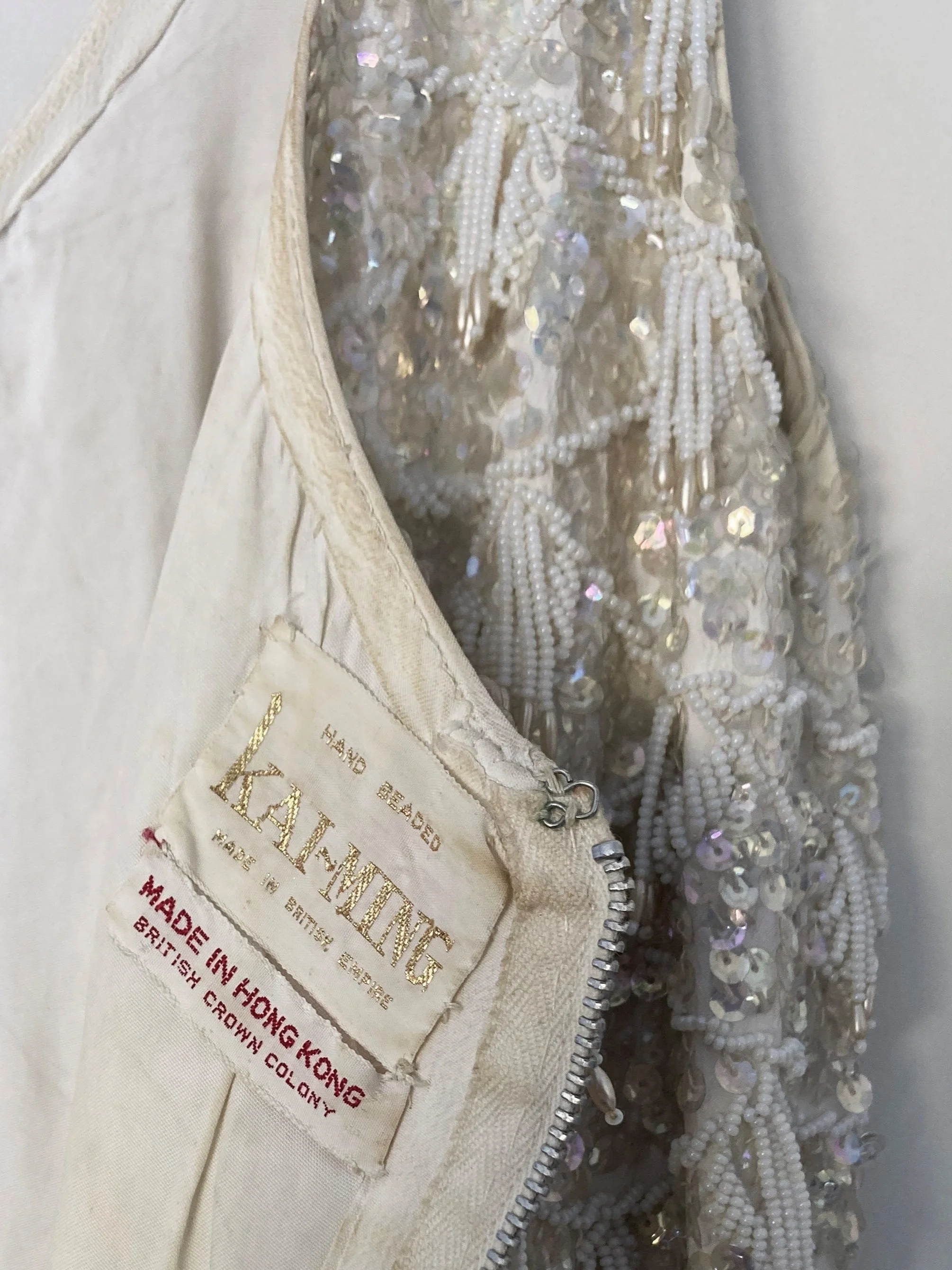 Close up view of the lining, label, metal zip closure and hook and eye on the hand beaded ivory evening gown. The label shows "Hand Beaded, Kai-Ming, Made in Hong Kong".