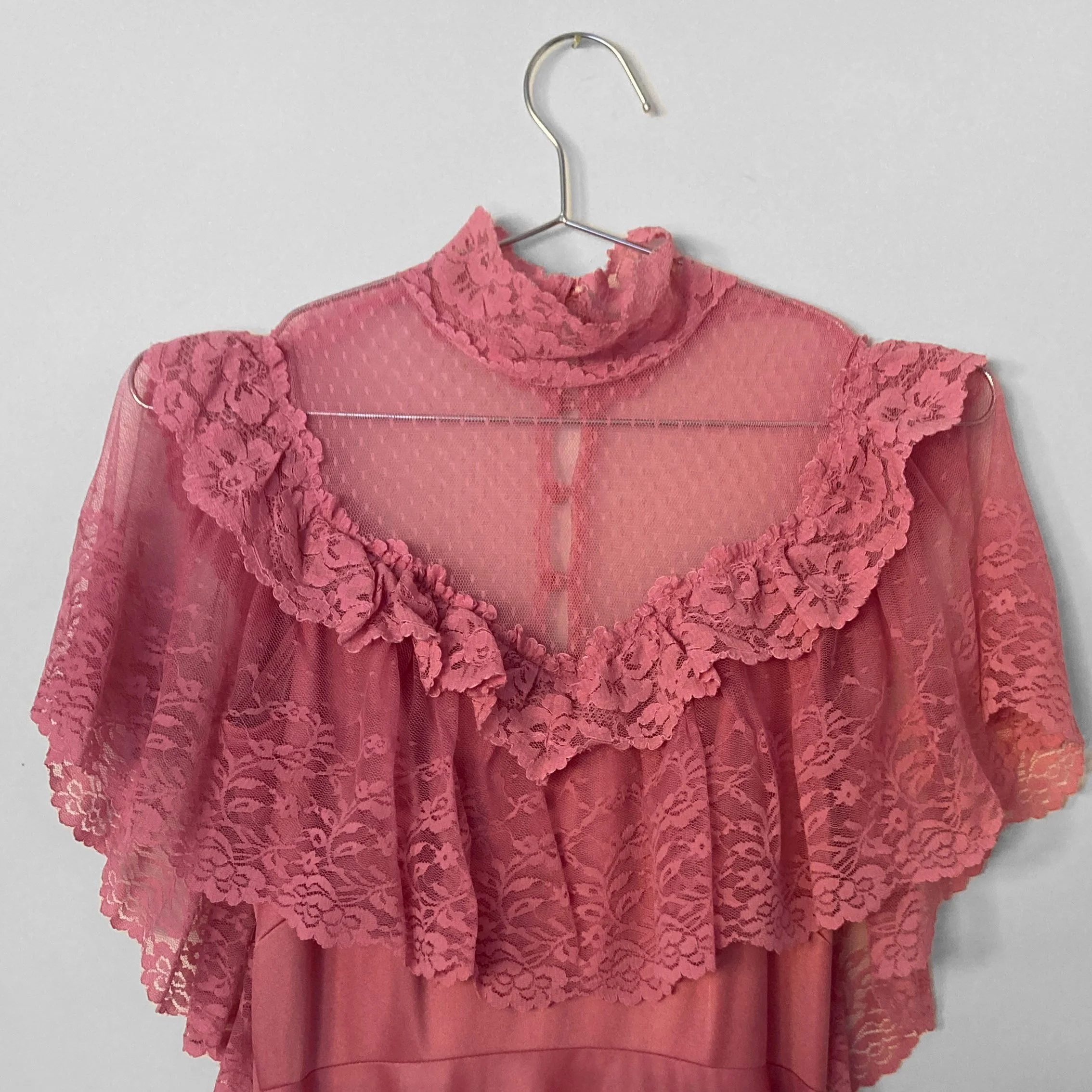 Close up image of the sheer lace bodice of a vintage rose pink gown. It highlights the lace neck, sheer décolletage and ruffles covering the bust.