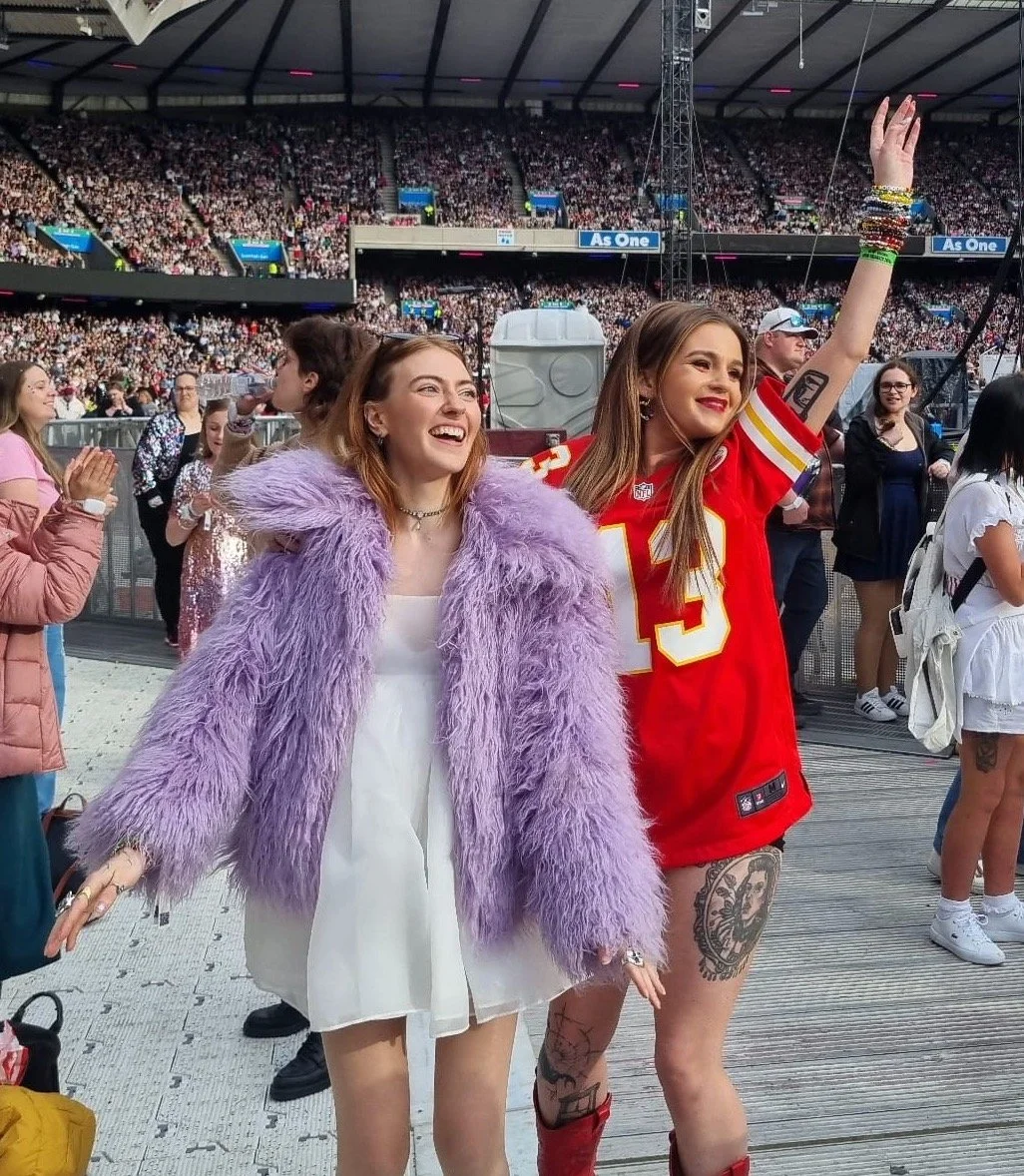 Two girls dance at the Eras Tour, one where's a lilac faux fur jacket over a white puff dress, the other wears a red "13" football jersey.