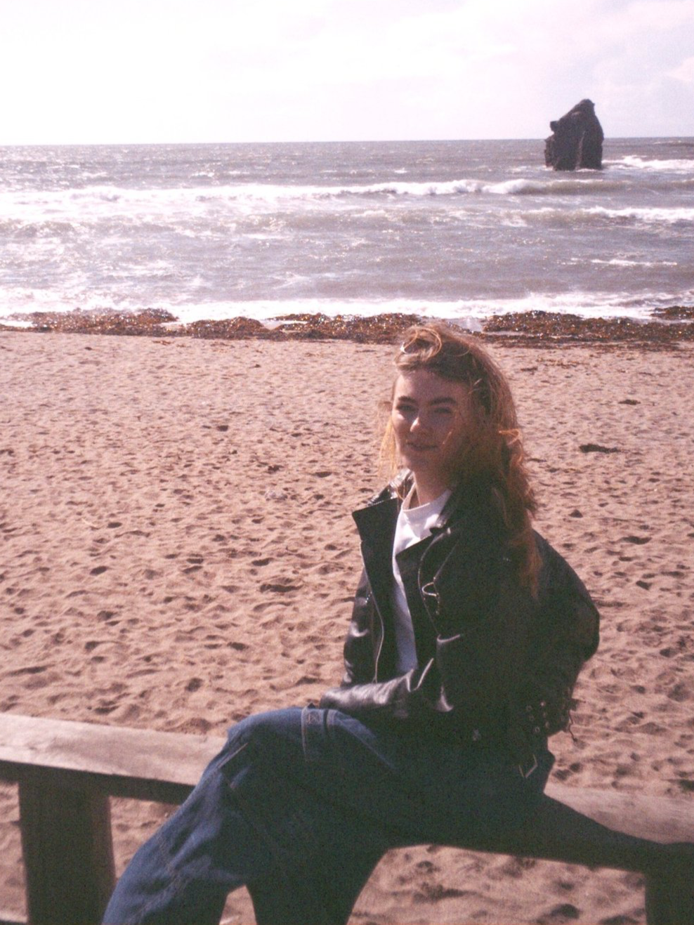 Phoebe Niemira sits on a beach in Devon wearing a vintage leather biker style jacket, white t-shirt and baggy jeans. Her sunglasses are on her head, pushing her messy windswept hair back off her face.