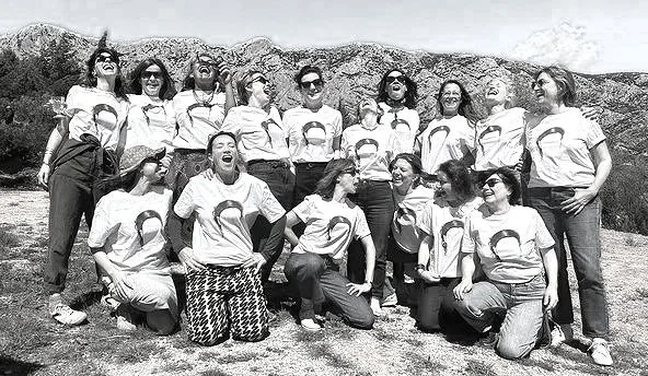 A group of women outdoors, standing and sitting, smiling and laughing, wearing casual clothing and sunglasses, with rocks and sky in the background.