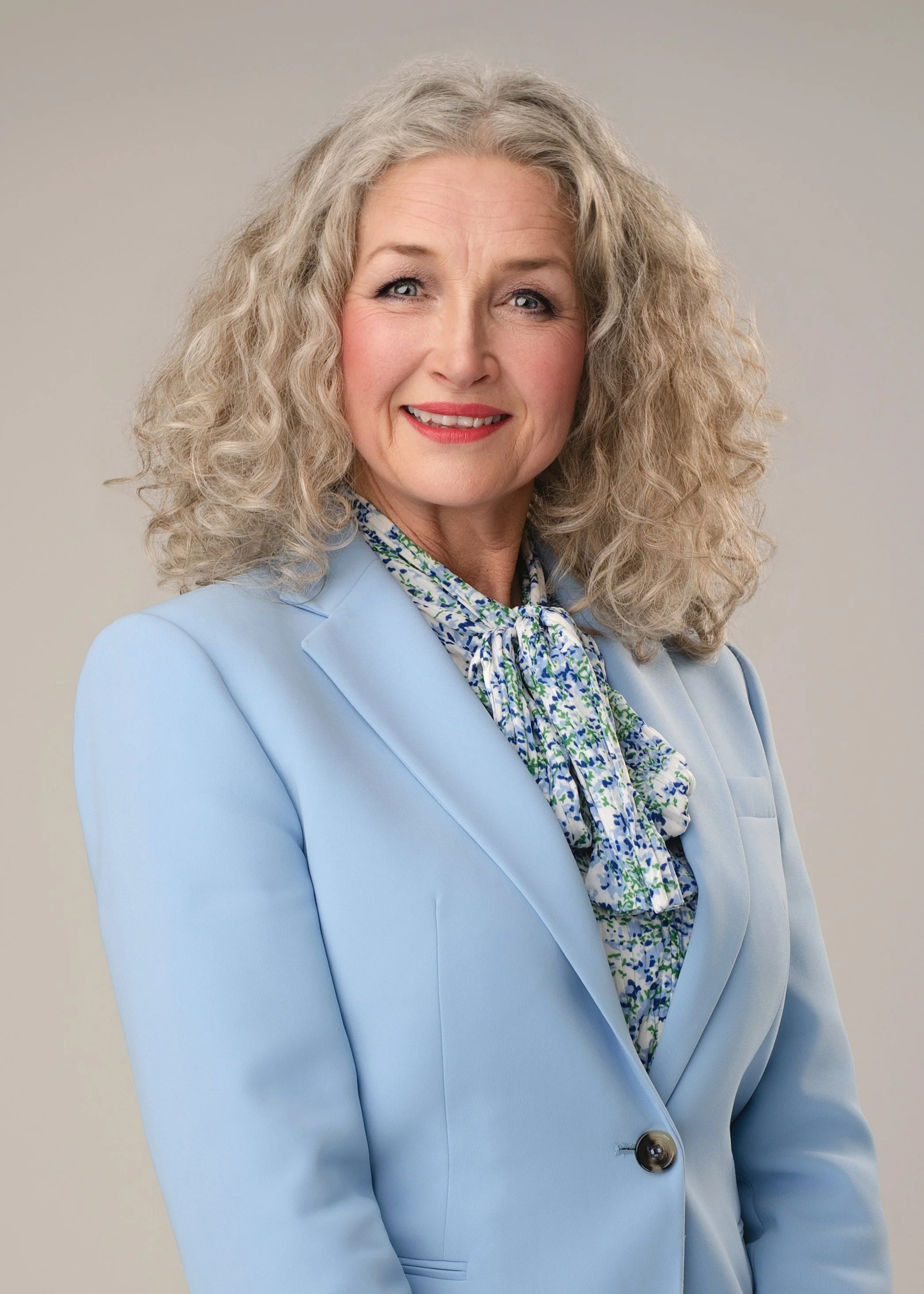 A business portrait of a woman with curly gray hair, blue eyes, and a warm smile, wearing a light blue blazer and a patterned scarf, against a plain gray background.