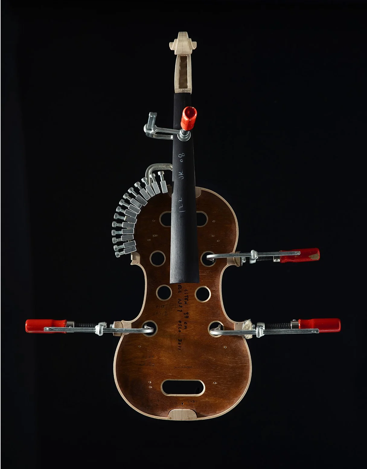 A wooden violin body with clamps, metal parts, and a fingerboard laid on top, set against a black background.