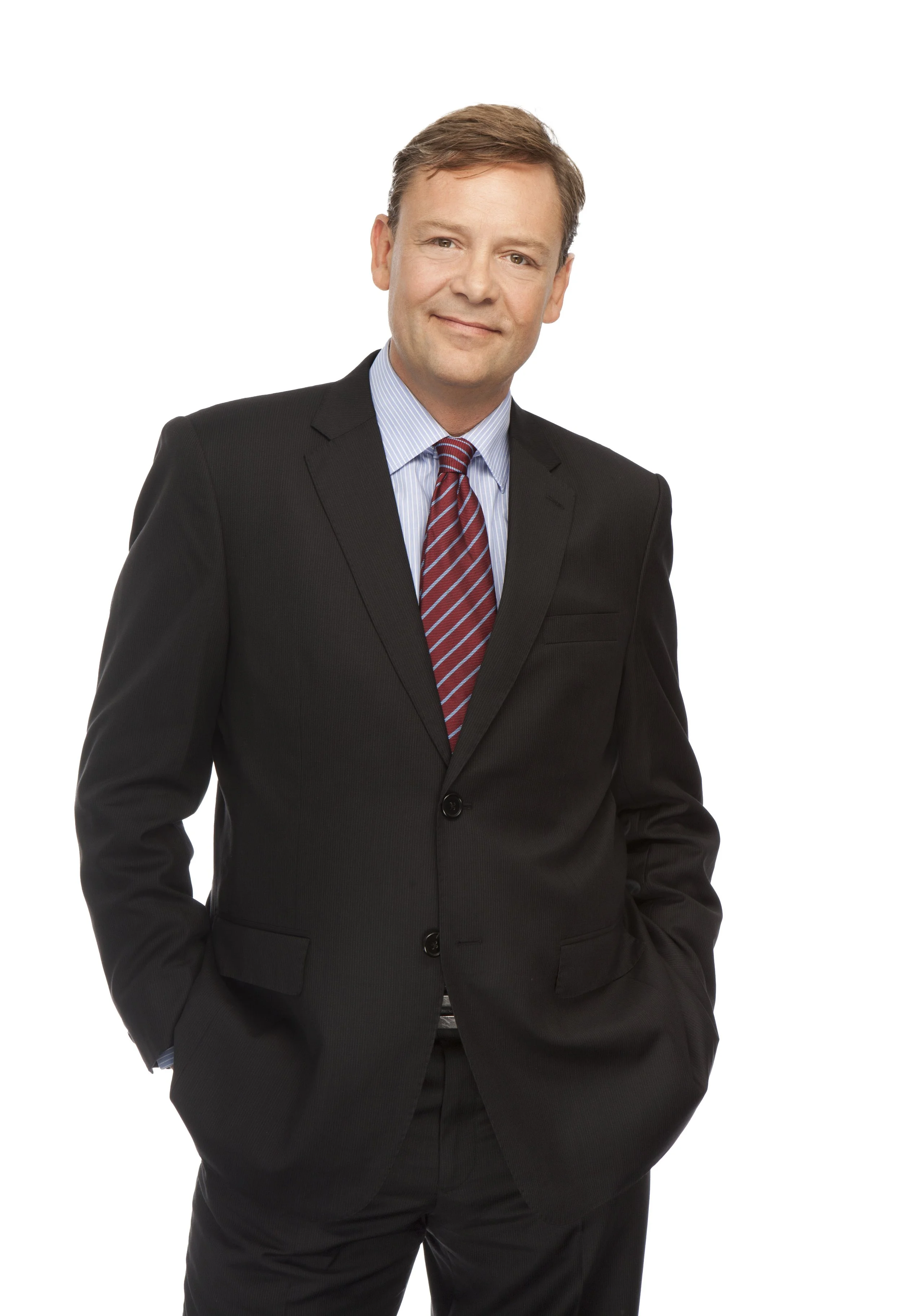 A professional man in a dark suit, blue striped shirt, and red striped tie, standing with one hand in his pocket, smiling at the camera against a white background.