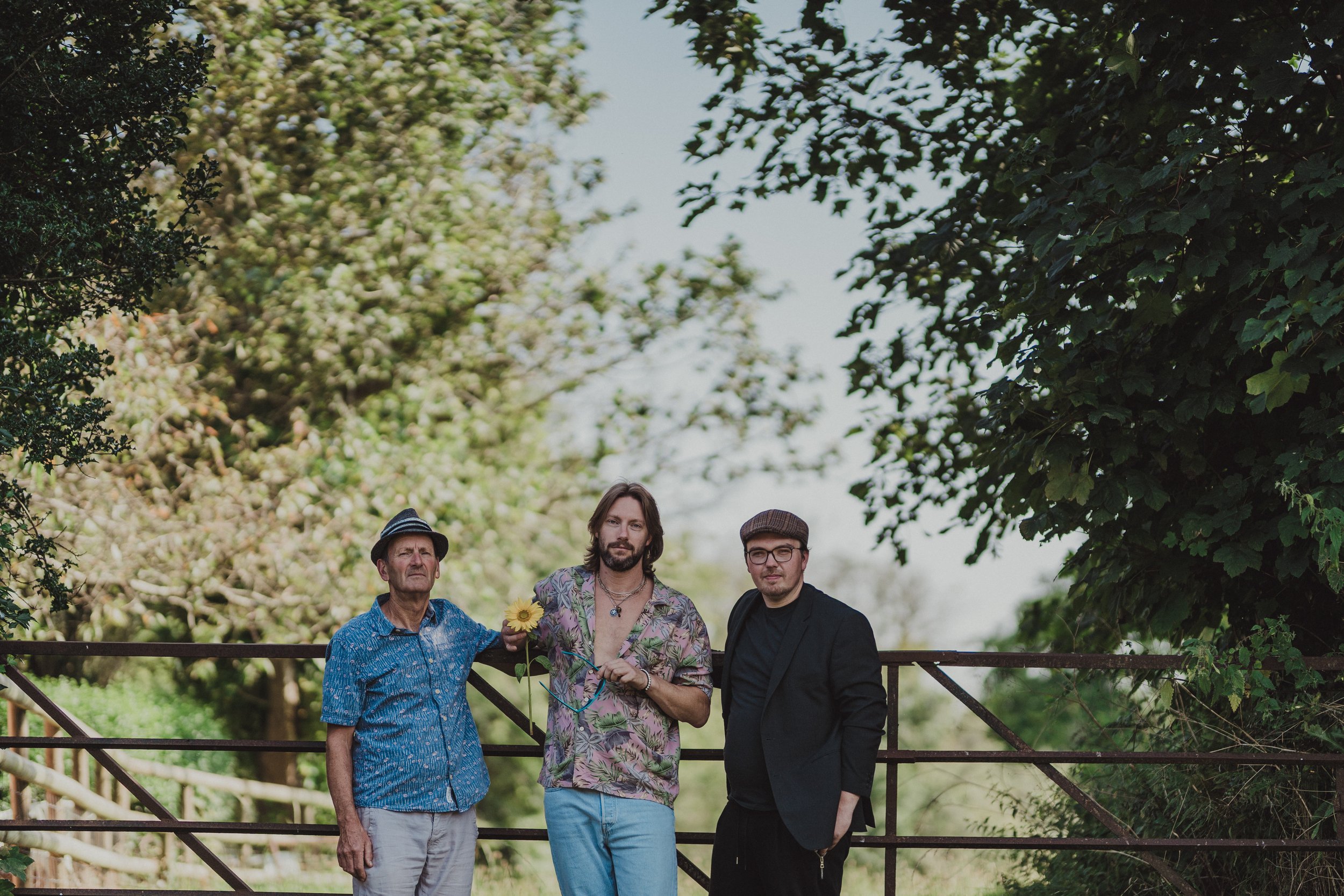 Three men standing together outdoors near a metal gate, surrounded by trees and greenery. One man is holding a sunflower and wearing a blue shirt and hat, the second is with long hair and a floral shirt, and the third man is wearing glasses, a hat, a