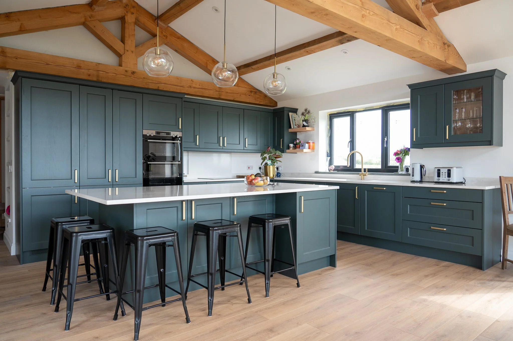 Modern kitchen with green cabinetry, white countertops, black barstools, wood ceiling beams, and a window above the sink. Cwtch Haus Kitchens.