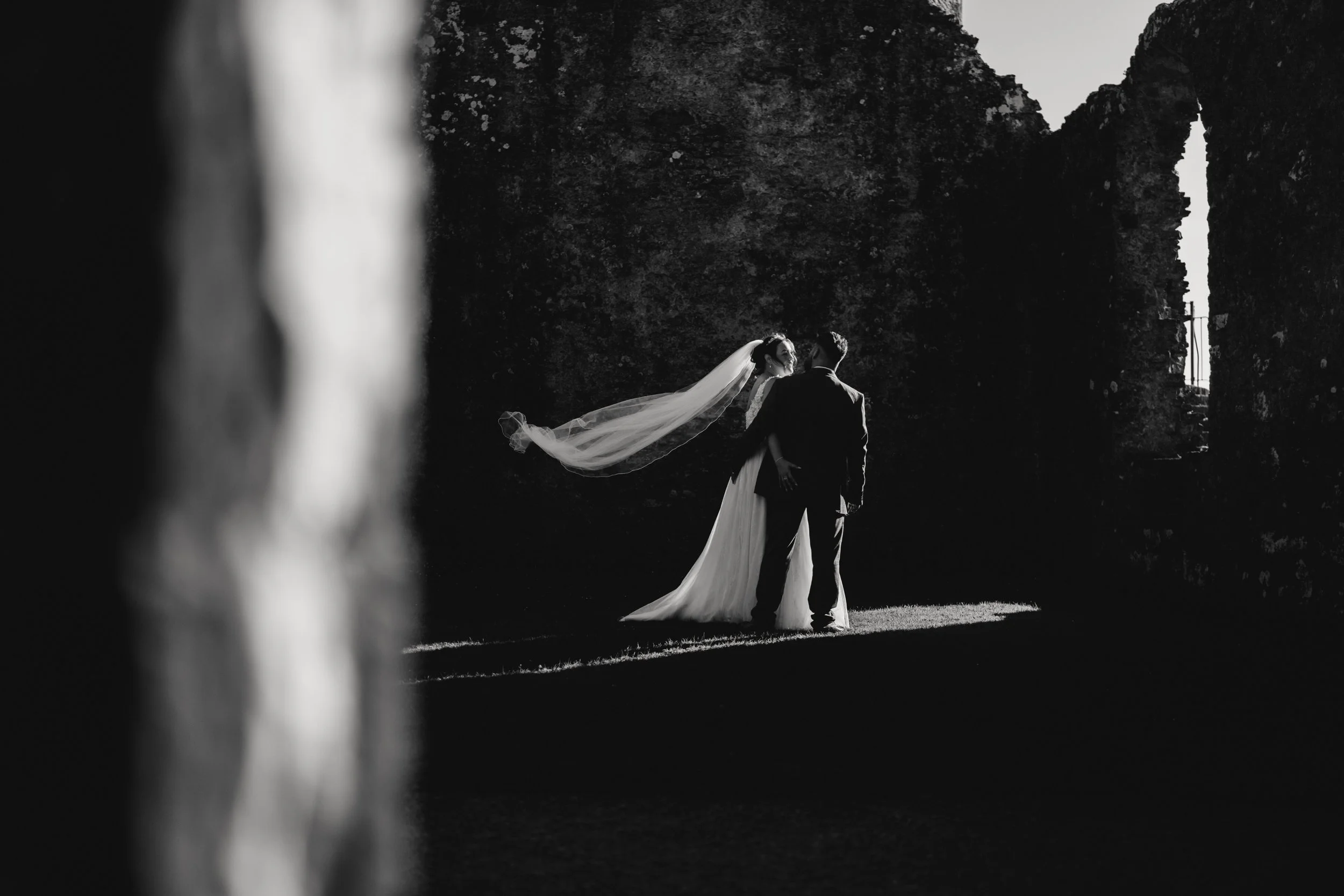 Black and white photo of a bride and groom standing outdoors, facing each other, with their faces close. The bride's veil is flowing in the wind. The background features dark trees and an old stone archway with a small bridge. The couple is illuminat
