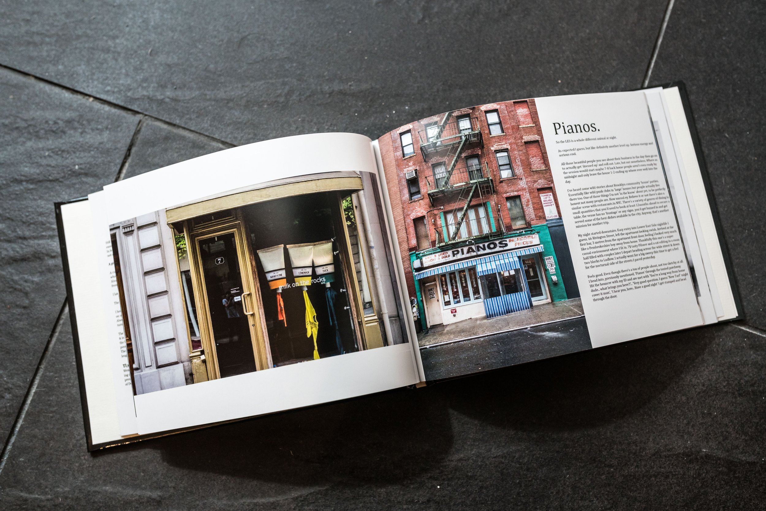 An open magazine on a dark tiled surface, showing photos of storefronts, including one with a sign that says 'Pianos' in New York and Paris and a building with a fire escape.
