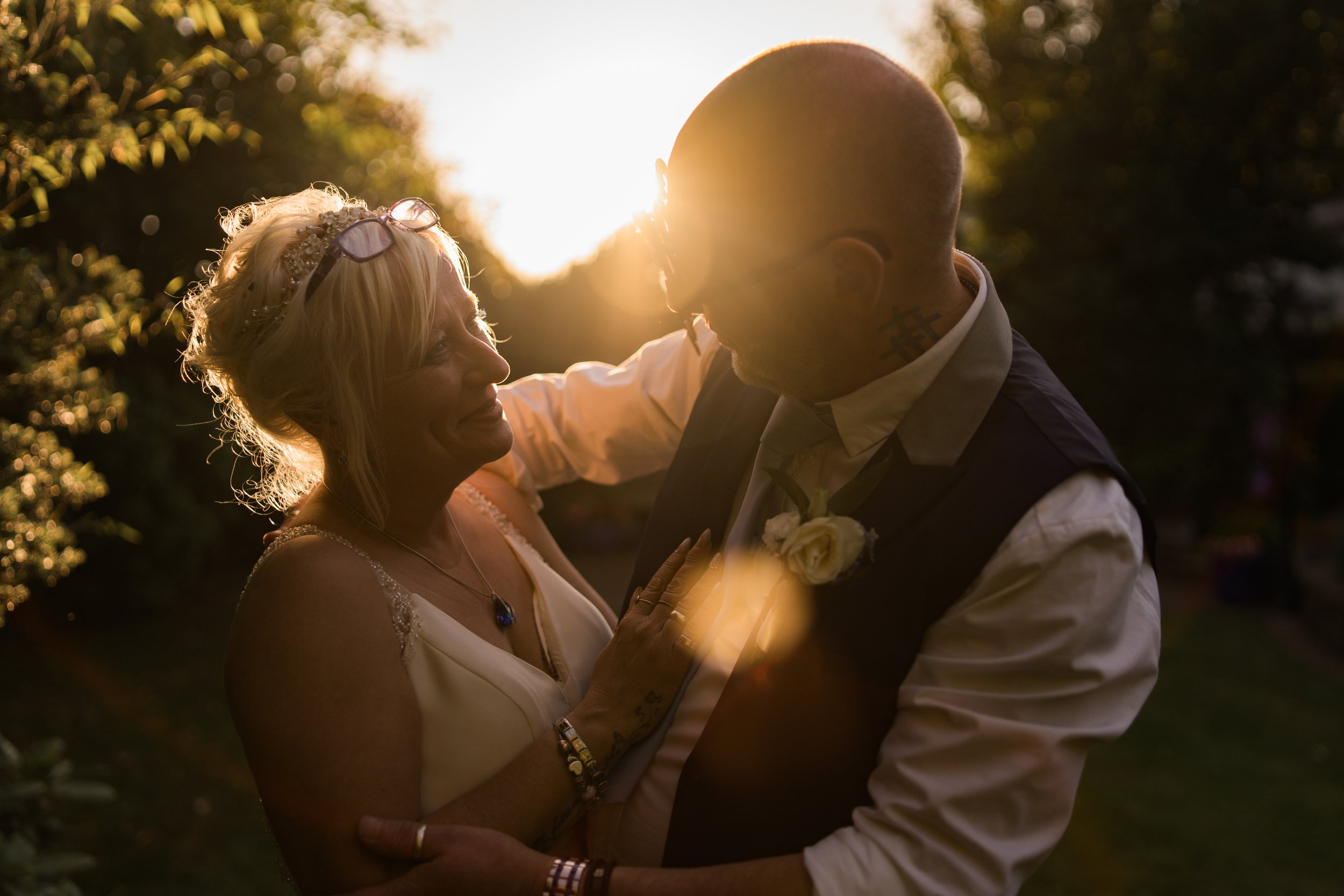 A middle-aged woman and a man with tattoos are dancing together outdoors during sunset. The woman has blonde hair, is wearing glasses on her head, a white dress, and jewelry. The man is bald, with facial tattoos, dressed in a vest and white shirt, wi