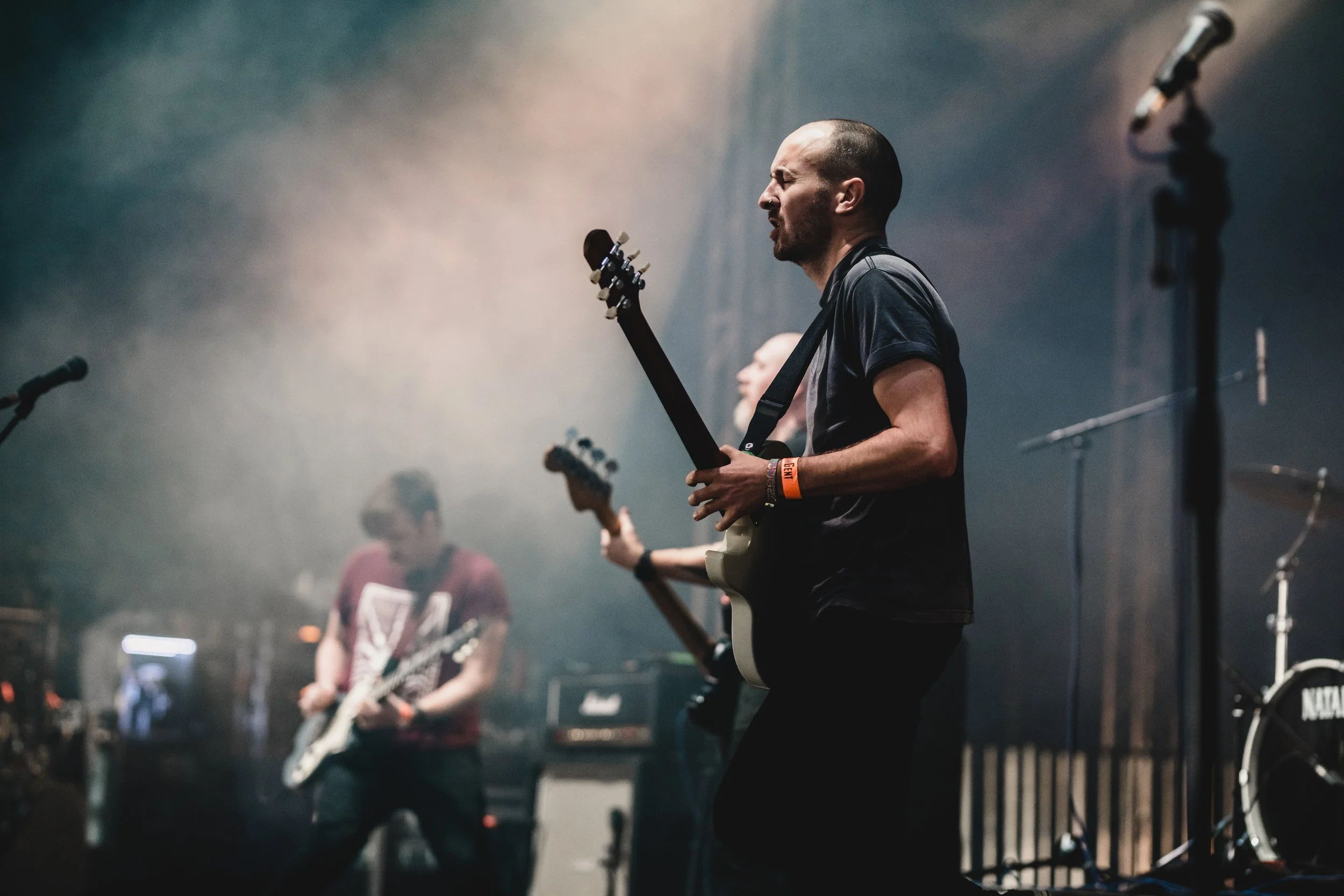 Musicians perform on stage with guitars and drums, surrounded by fog and stage equipment.