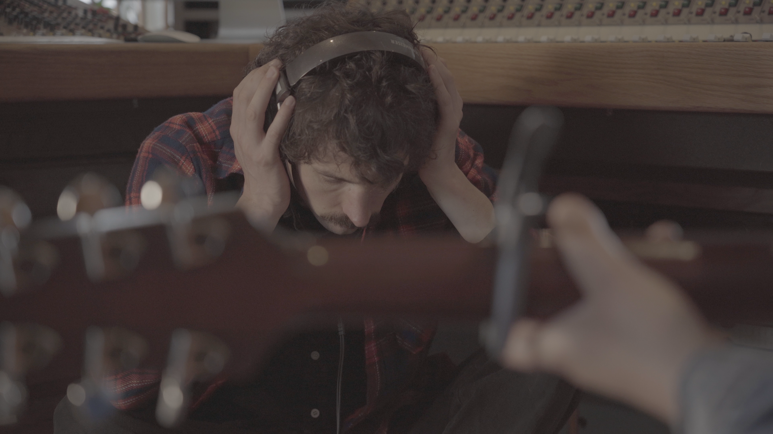 A man with curly hair and a plaid shirt, crouching with his head down, holding headphones to his ears, in a recording studio or music space.