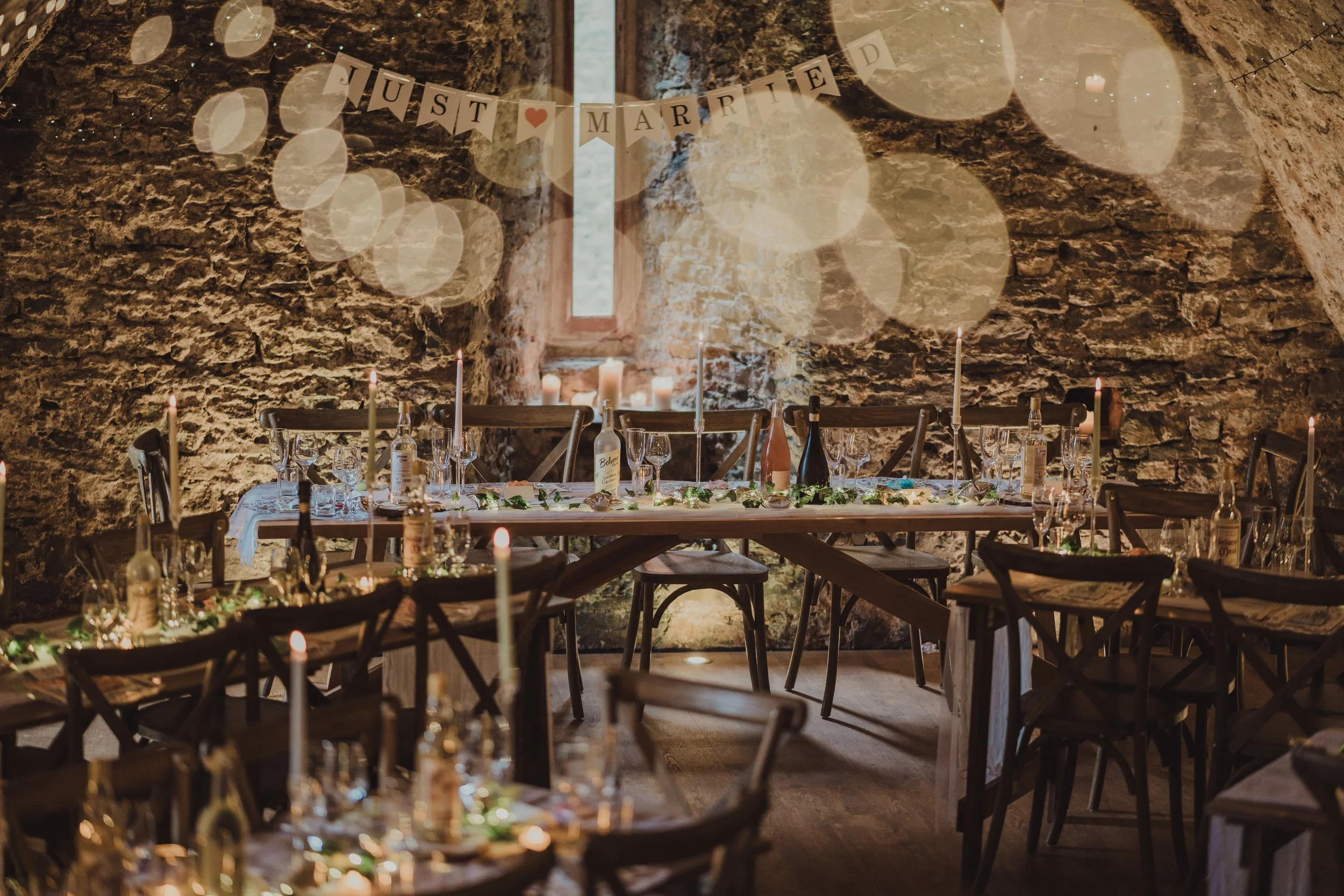 Indoor wedding reception with rustic stone walls, a banner reading 'Just Married' in the background, and a long table decorated with candles, bottles, and greenery. Alt Wedding. Alternative wedding photography.