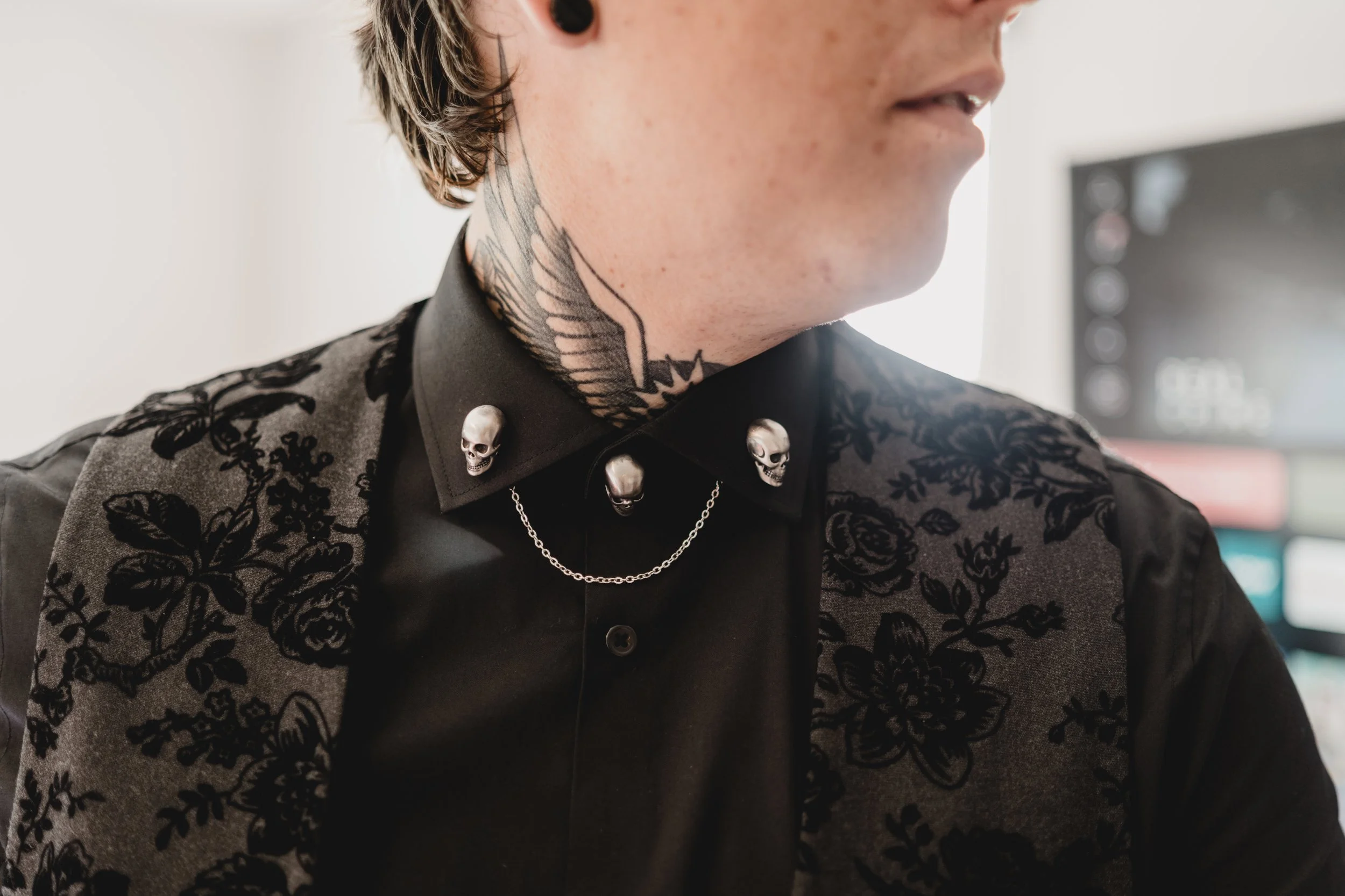 Close-up of a person's neck and shoulder area with tattoos and gothic-style clothing, including a black shirt with skull and chain details and a floral-patterned shoulder piece.