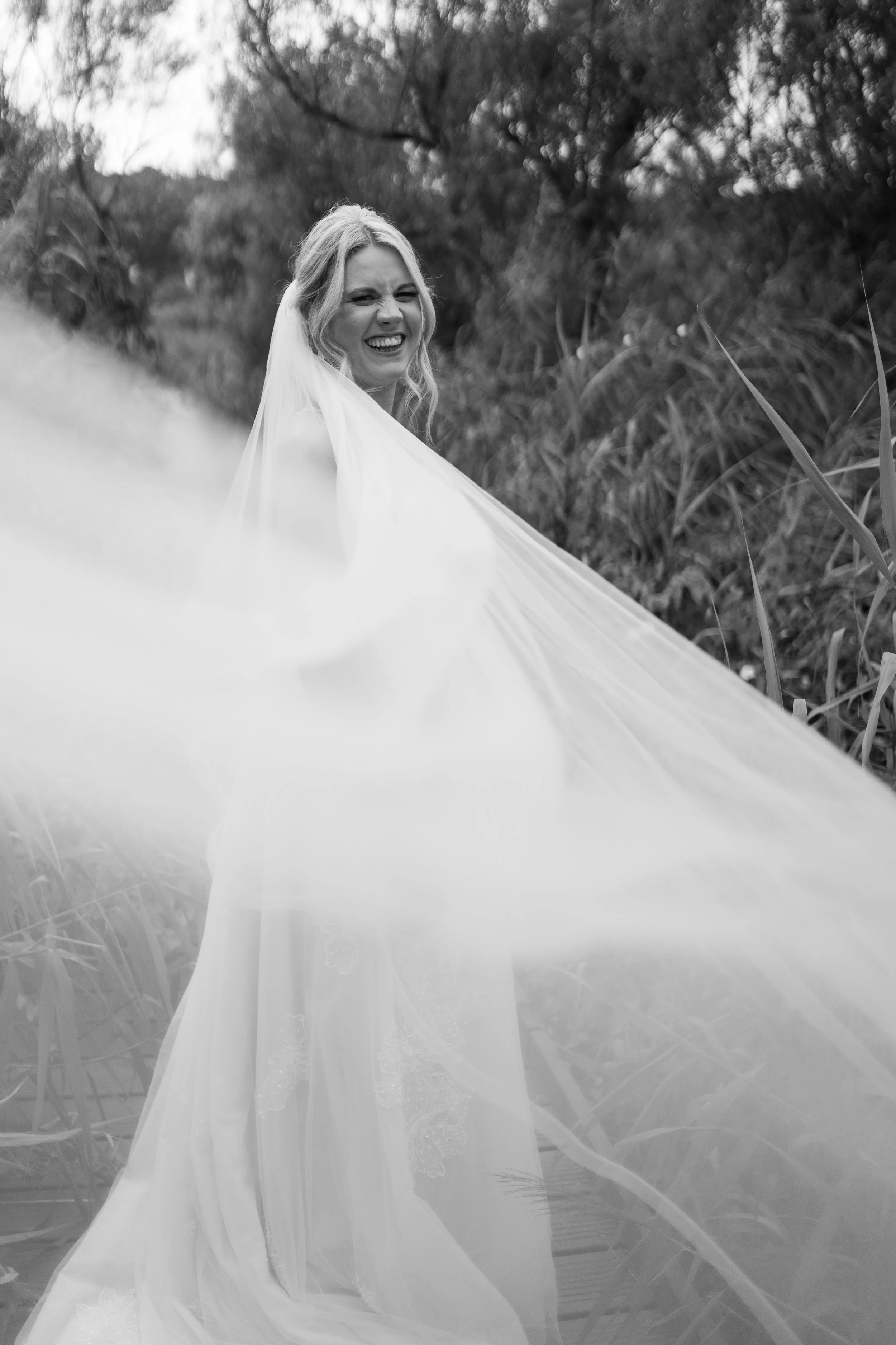 A smiling bride in a wedding dress and veil stands outdoors among tall grass and trees.