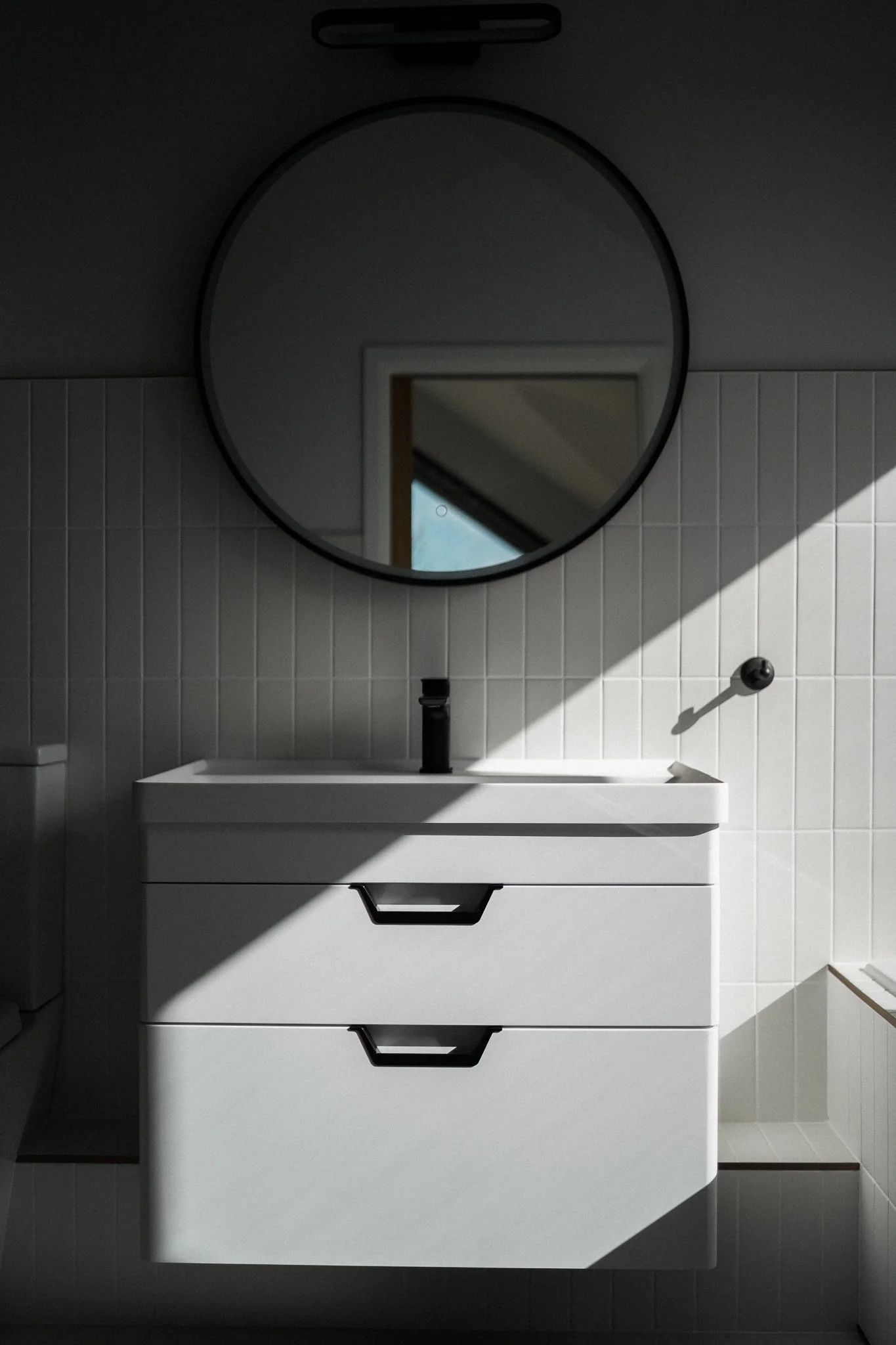 Minimalist bathroom with white vanity, black faucet, round mirror, and tiled wall in natural sunlight.