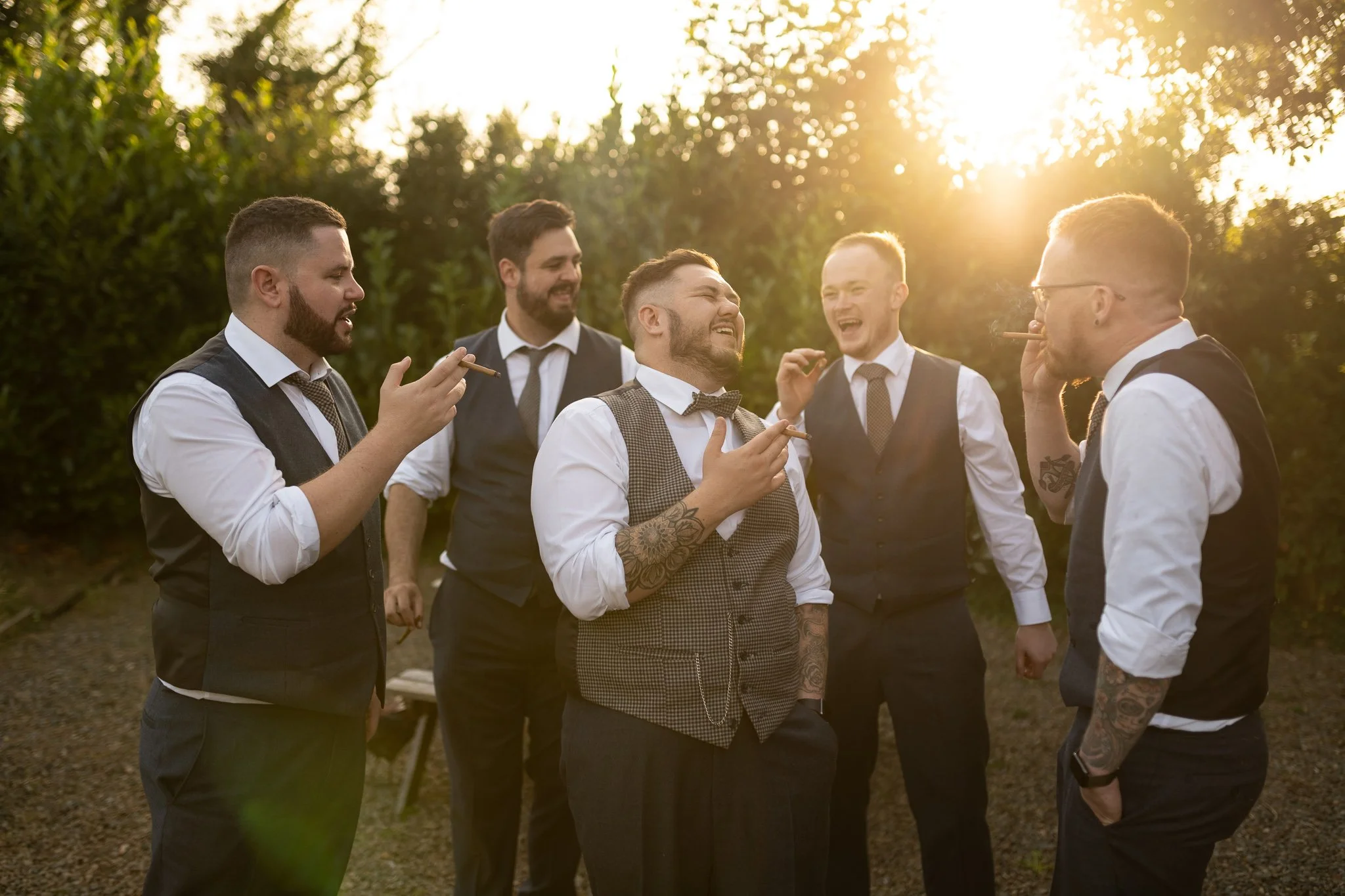 A group of six men dressed in formal vests and shirts, standing outdoors during sunset, laughing and smoking cigars.