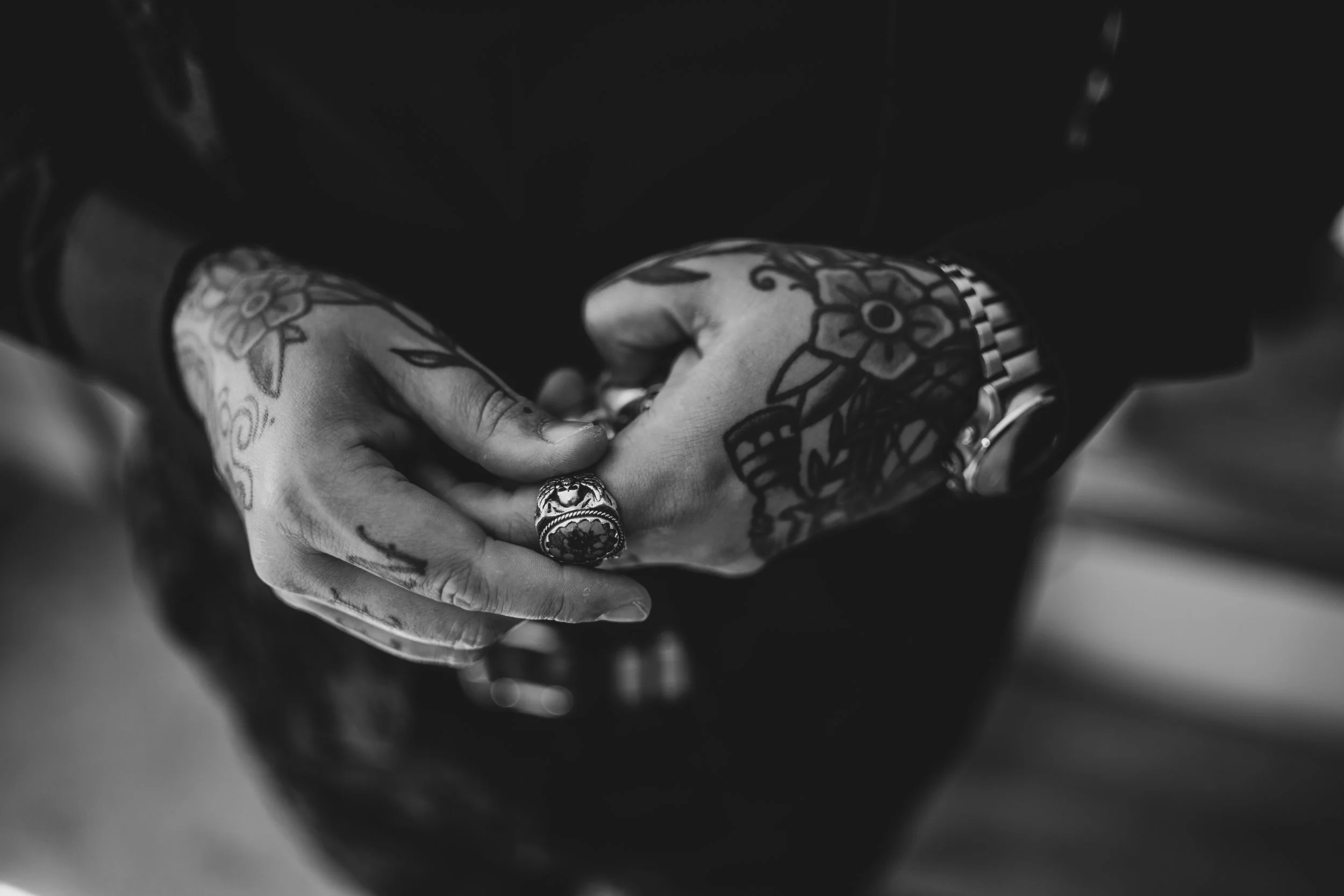 Close-up of tattooed hands with rings and a bracelet, clasped together.