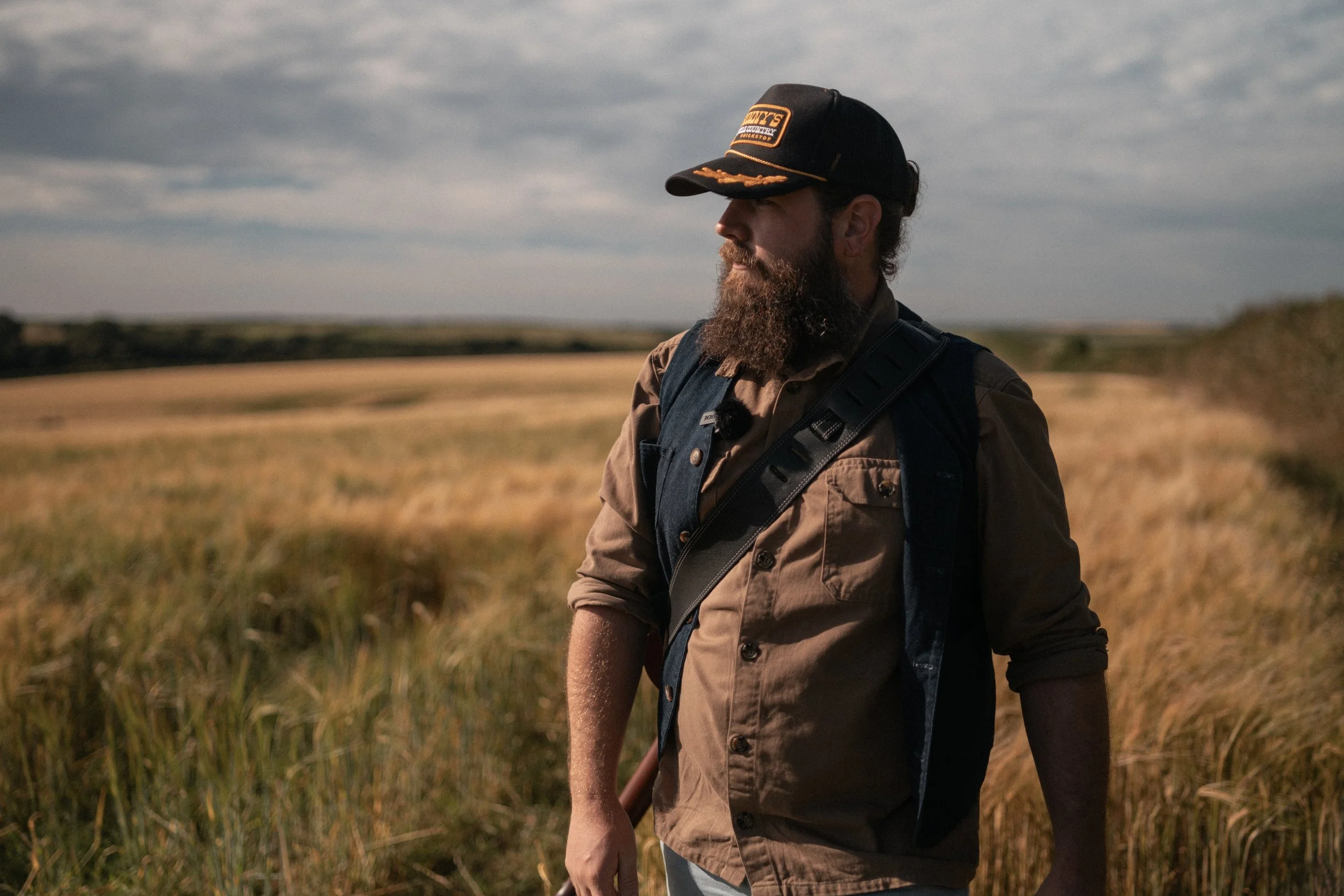 A man with a beard wearing a black cap and brown shirt, standing in a field of golden grass with a cloudy sky overhead. Promotional Music Photography.