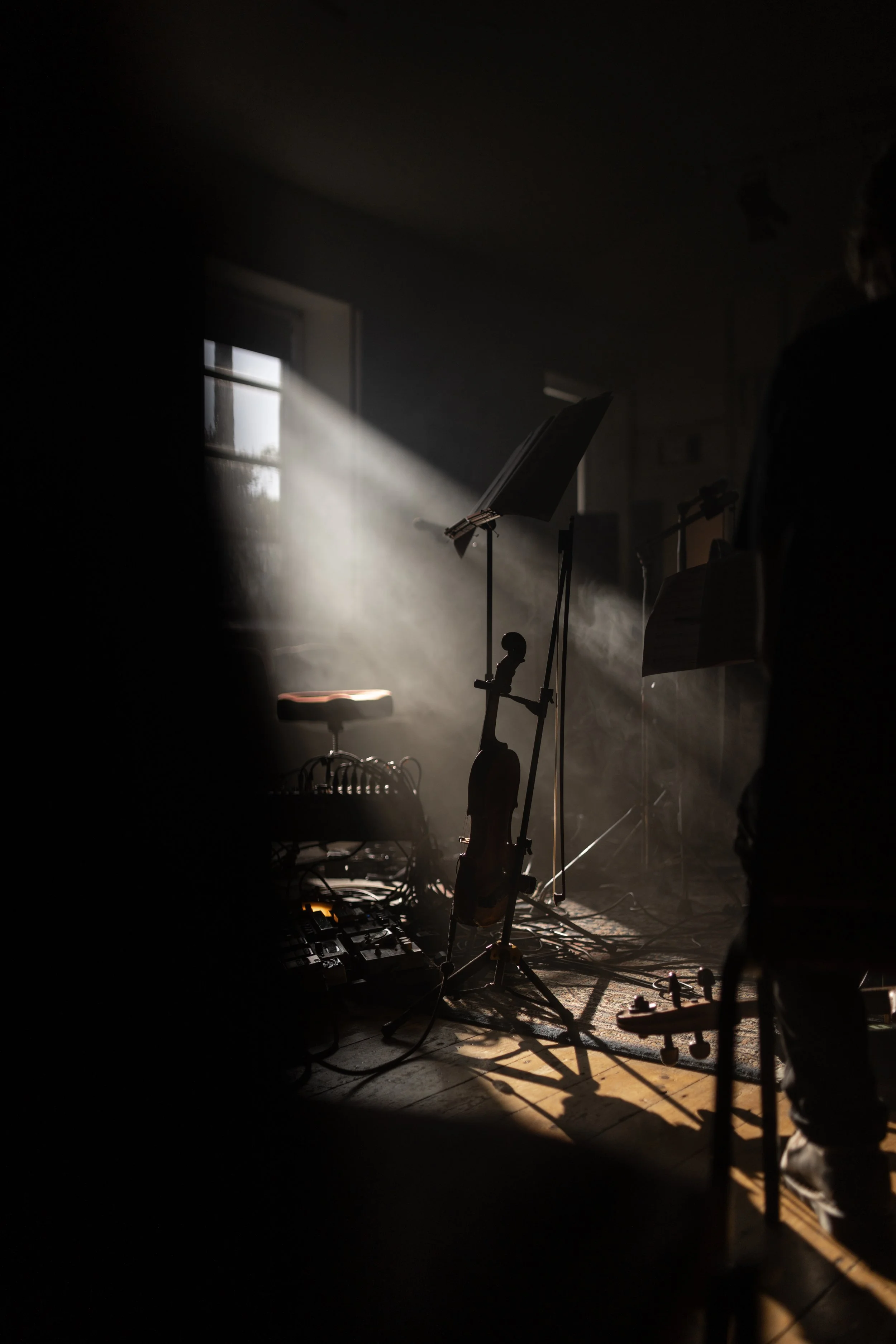 Musician's setup includes a violin on a stand, sheet music, a music stand, and other audio equipment near a window with sunlight streaming in. Live event photography.