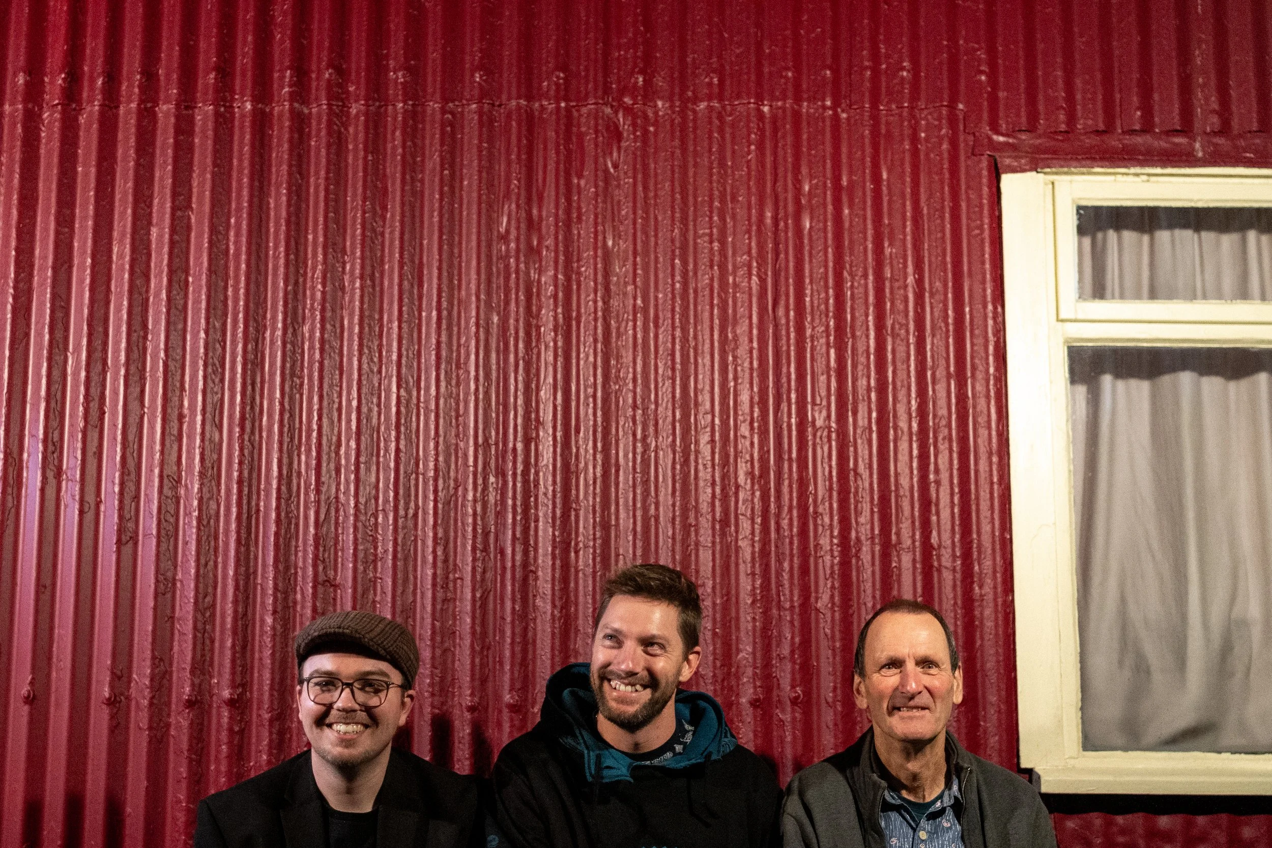 Three men standing in front of a red corrugated metal wall with a window on the right side. Promotional Music Photography.