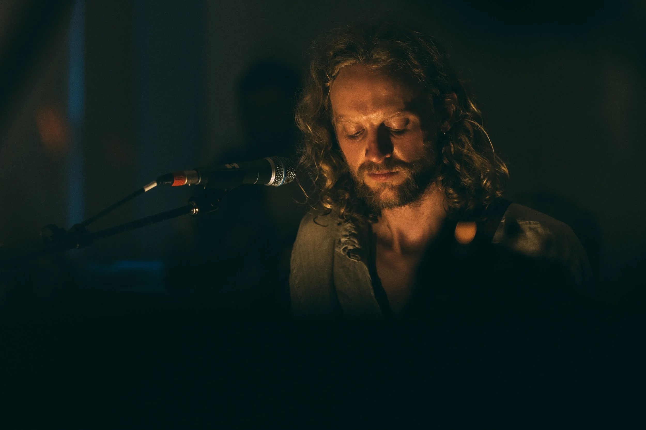 A man with long, curly hair and a beard, performing in front of a microphone in a dimly lit setting. Promotional Music Photography.