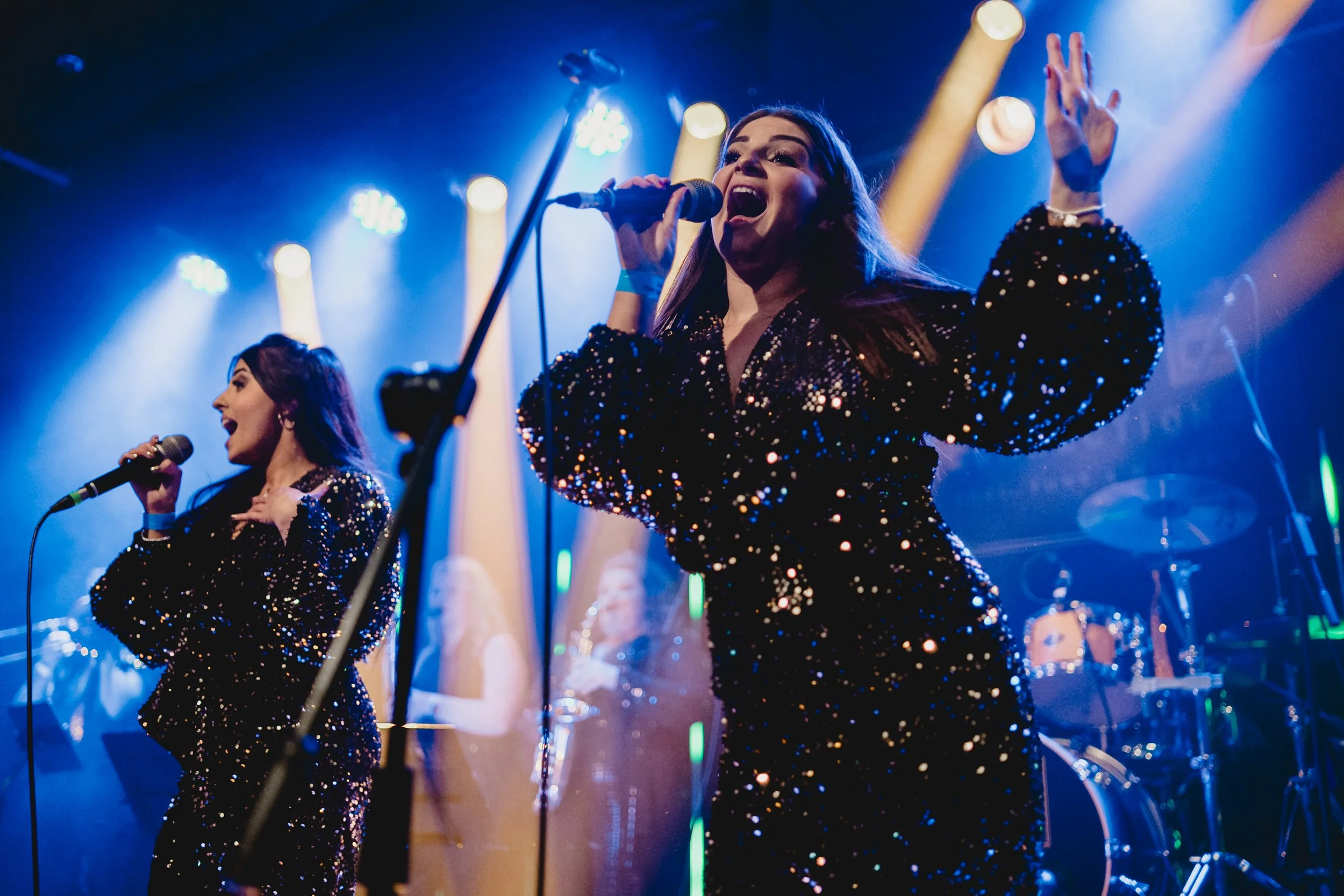 Two female singers performing on stage with colorful lighting, wearing matching black sequined outfits, with drums and musical equipment in the background. Live event photography.