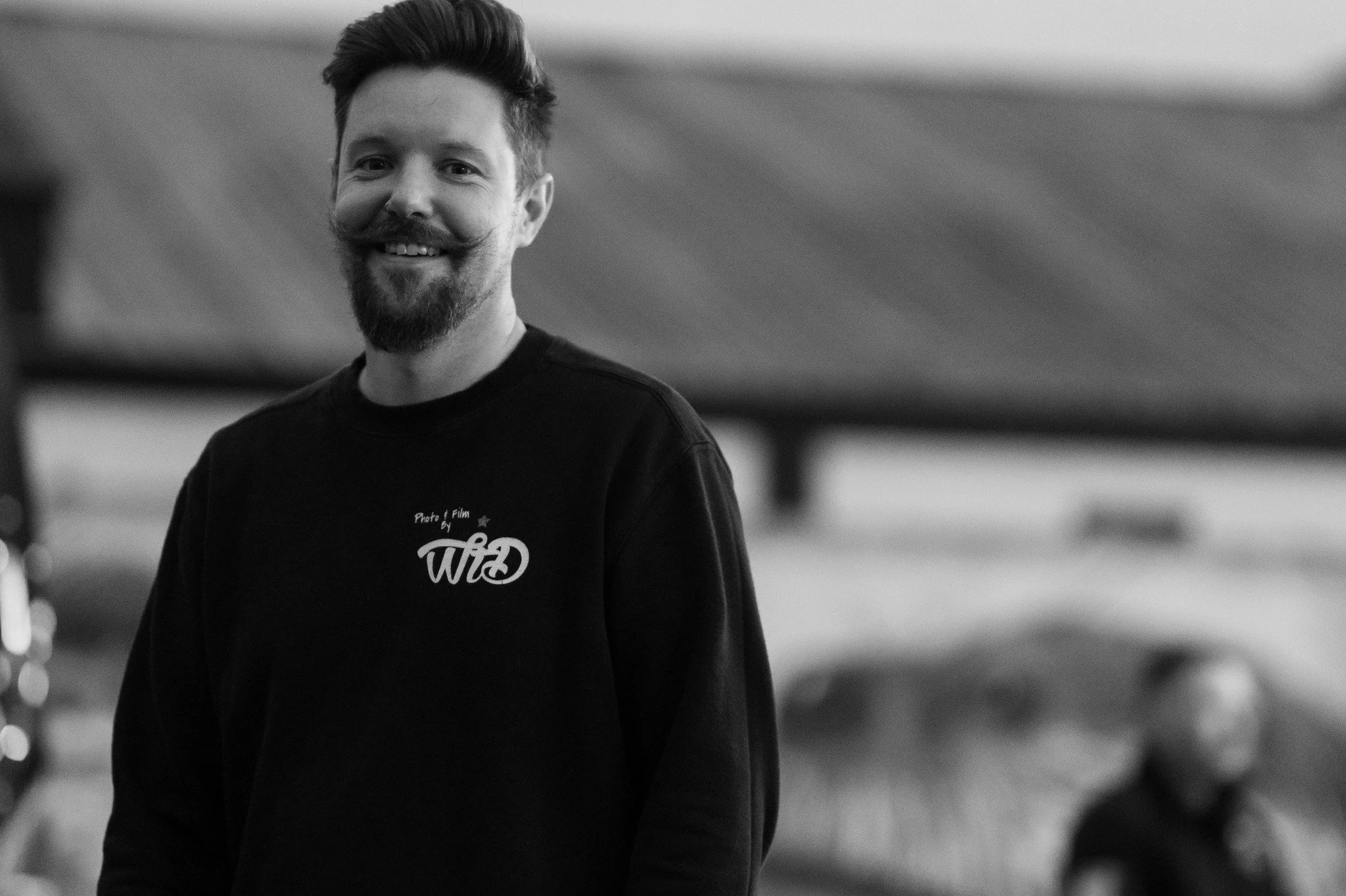 Black and white photo of a smiling man with a beard and styled hair, wearing a black sweatshirt with white text, standing outdoors with a blurred background.