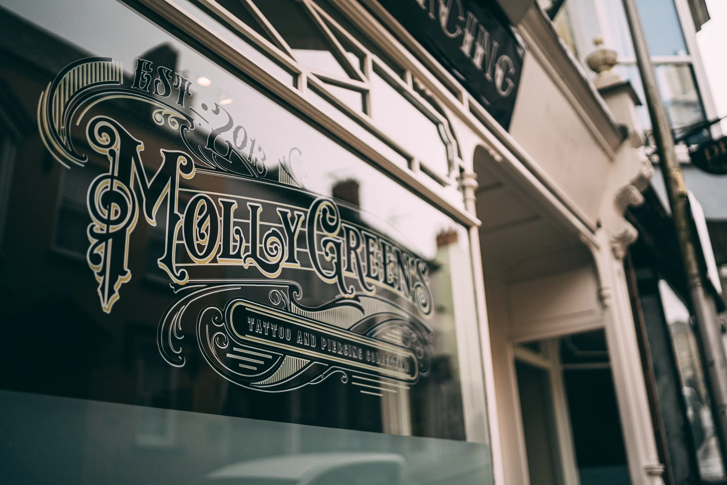 Close-up of a decorative glass storefront window with vintage-style gold lettering that reads "Established 2013 Moly Greens Tattoo and Piercing Collective." The storefront has ornate architectural details, and a reflection of buildings is visible on 