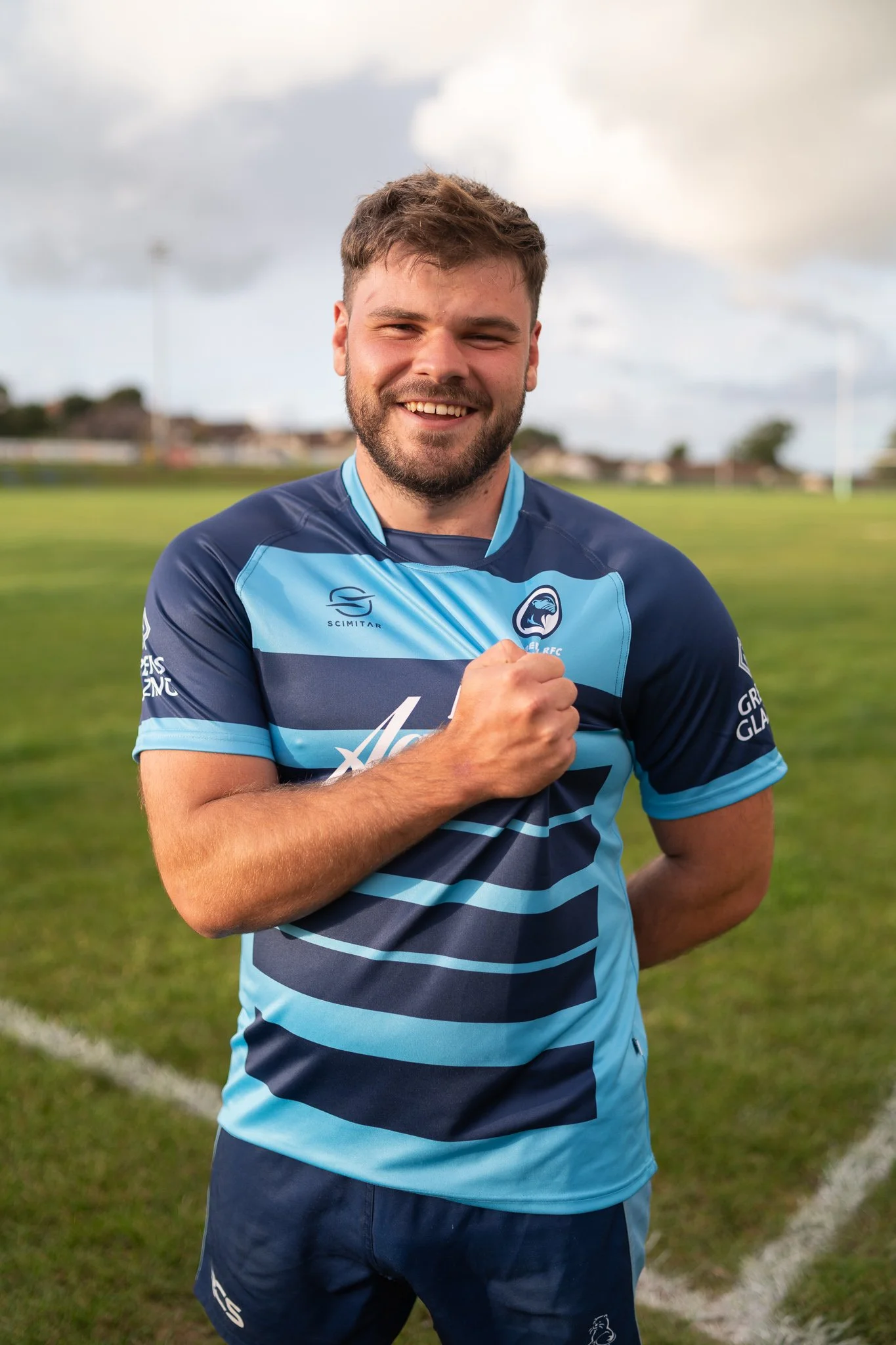 A young man with short brown hair and a beard, wearing a blue Rugby uniform, smiling on a grassy soccer field. Narberth RFC.