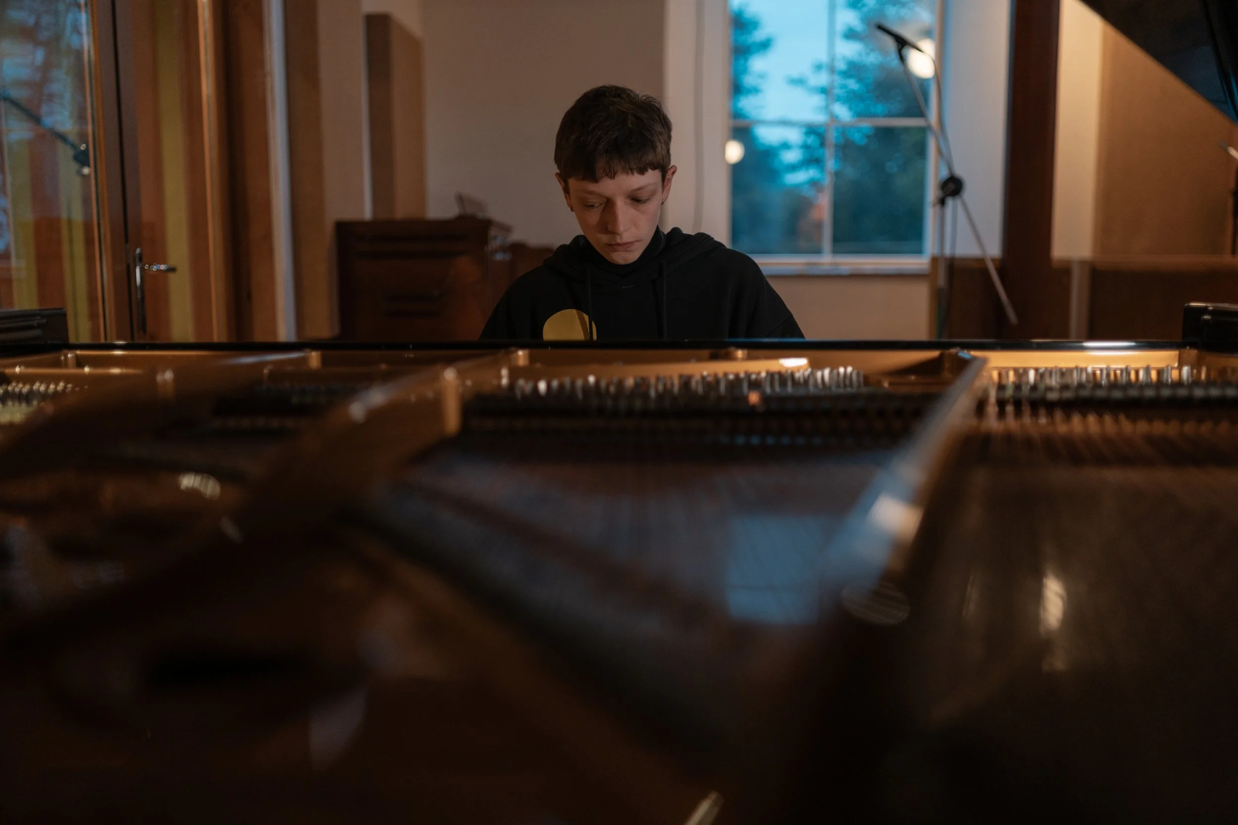 Young boy sitting in front of an open grand piano in a room with large windows and a blue sky outside.