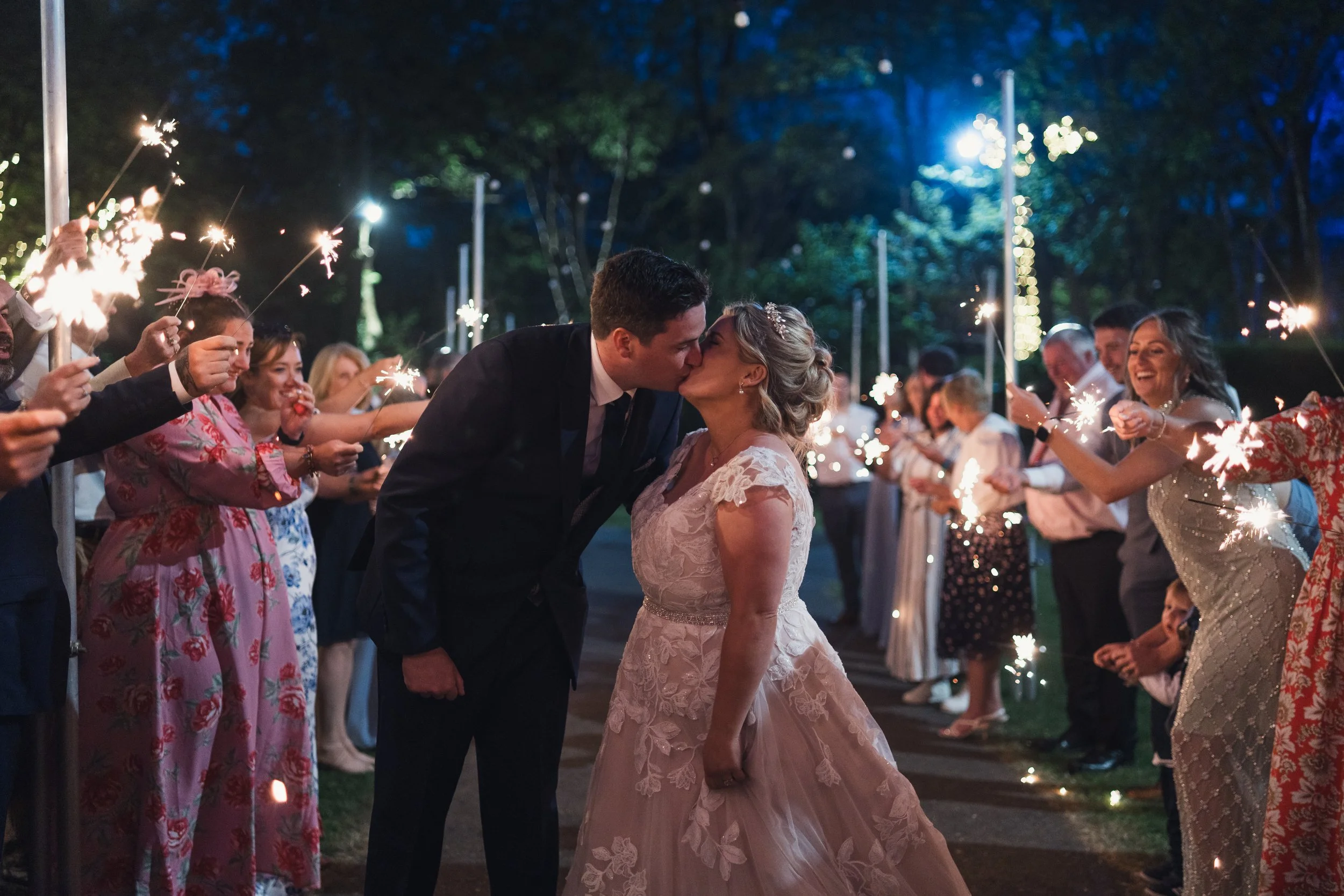 A newlywed couple kissing during a celebration surrounded by friends and family holding sparklers at night.