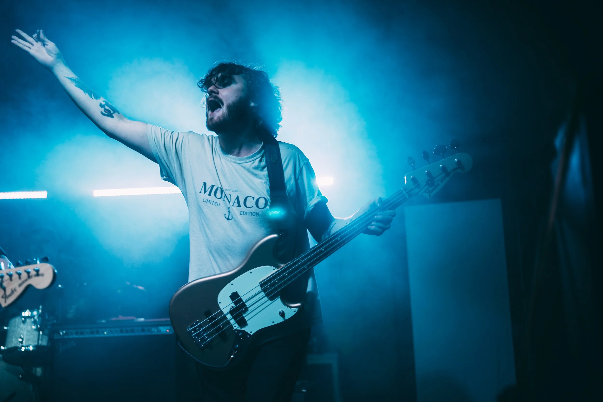 A male musician with curly hair and a beard singing and playing an electric bass guitar on stage, wearing a white T-shirt with the word 'MONACO' printed on it, illuminated by blue stage lighting. Live event Photography.