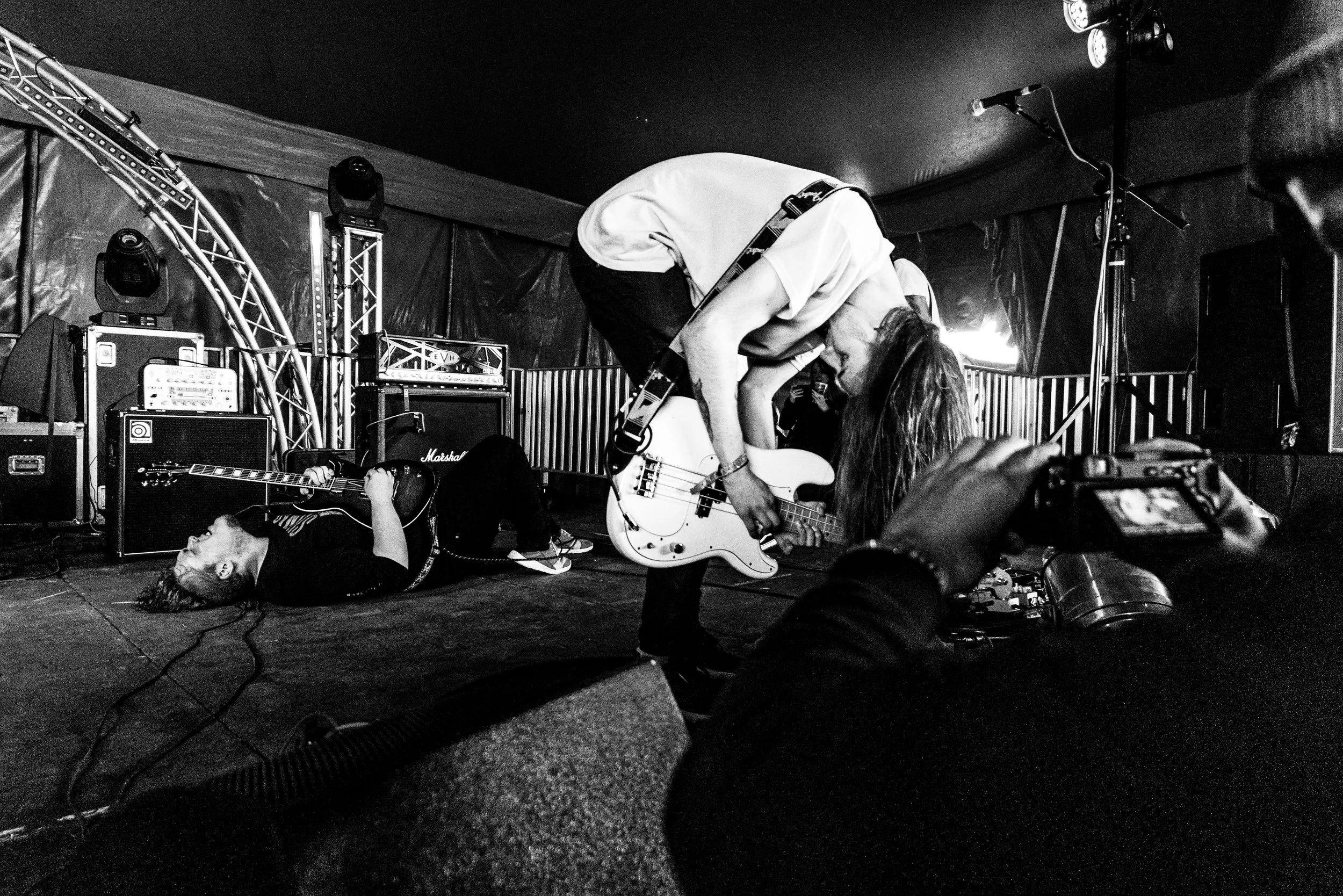 Black and white photo of a live music performance with two musicians: one playing a bass guitar and bent over with long hair obscuring their face, and the other lying on the floor playing an electric guitar, with a person taking a photo in the foregr