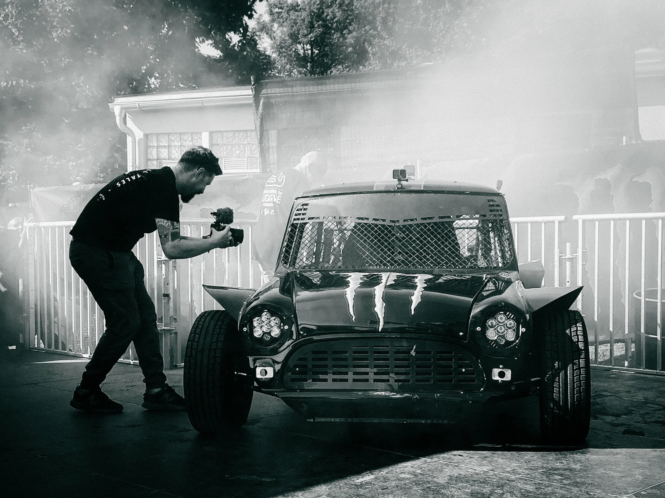A man is filming a race car with a camera, behind a fog or smoke effect, next to a race track fence.