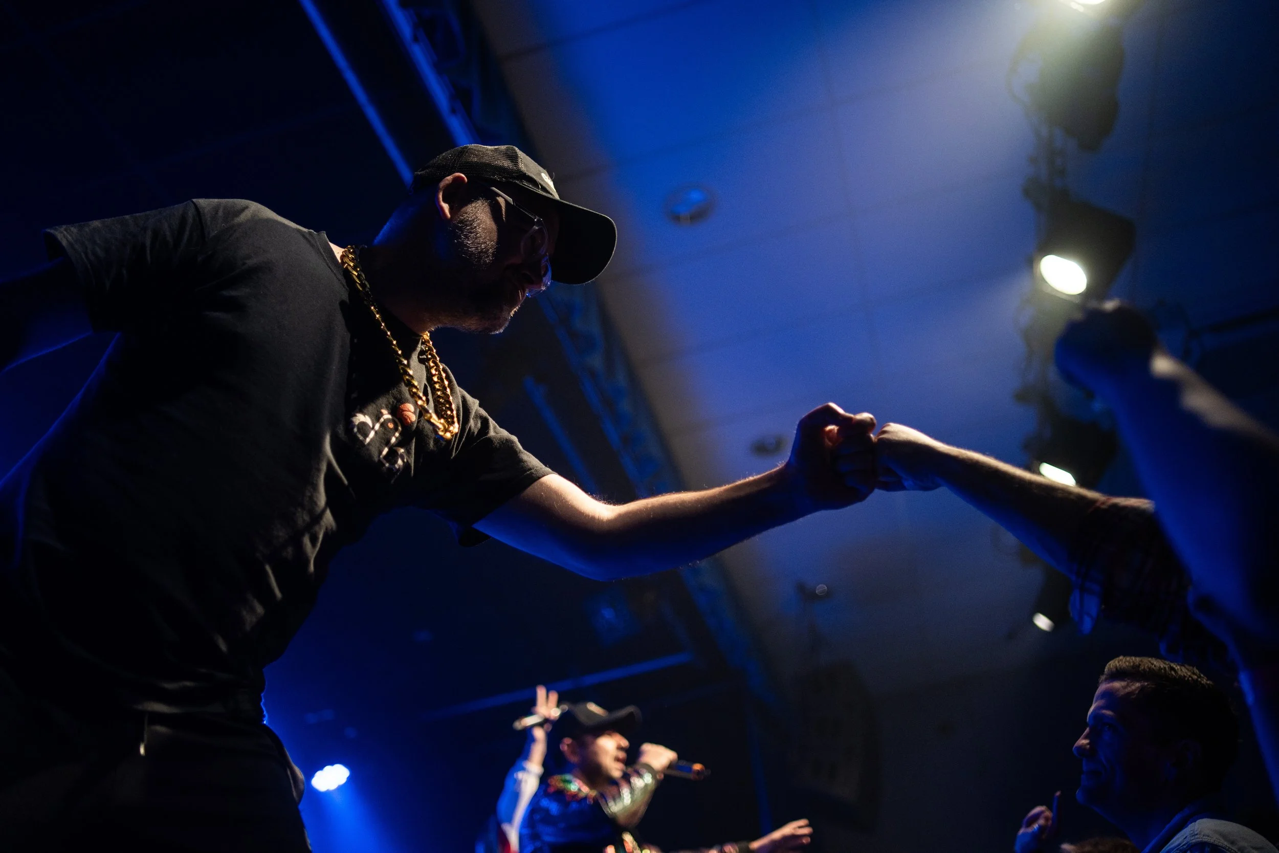 Person wearing a cap, sunglasses, and gold chains fist bumps another person in a dark, blue-lit crowd at a concert or event, with stage lights and a performer in the background.
