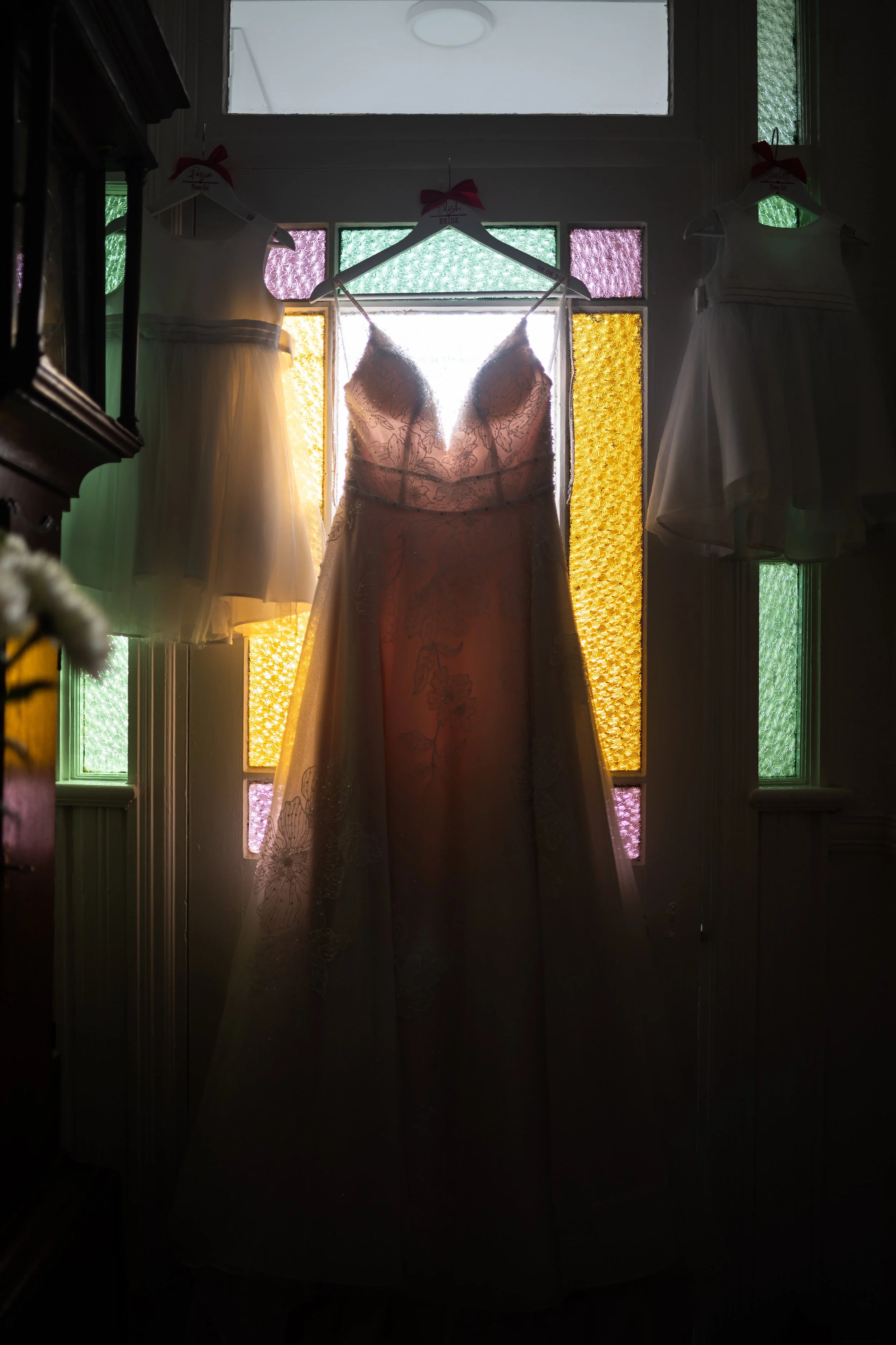 A wedding dress hanging in front of a stained glass window with colorful panels, backlit to create a silhouette effect.