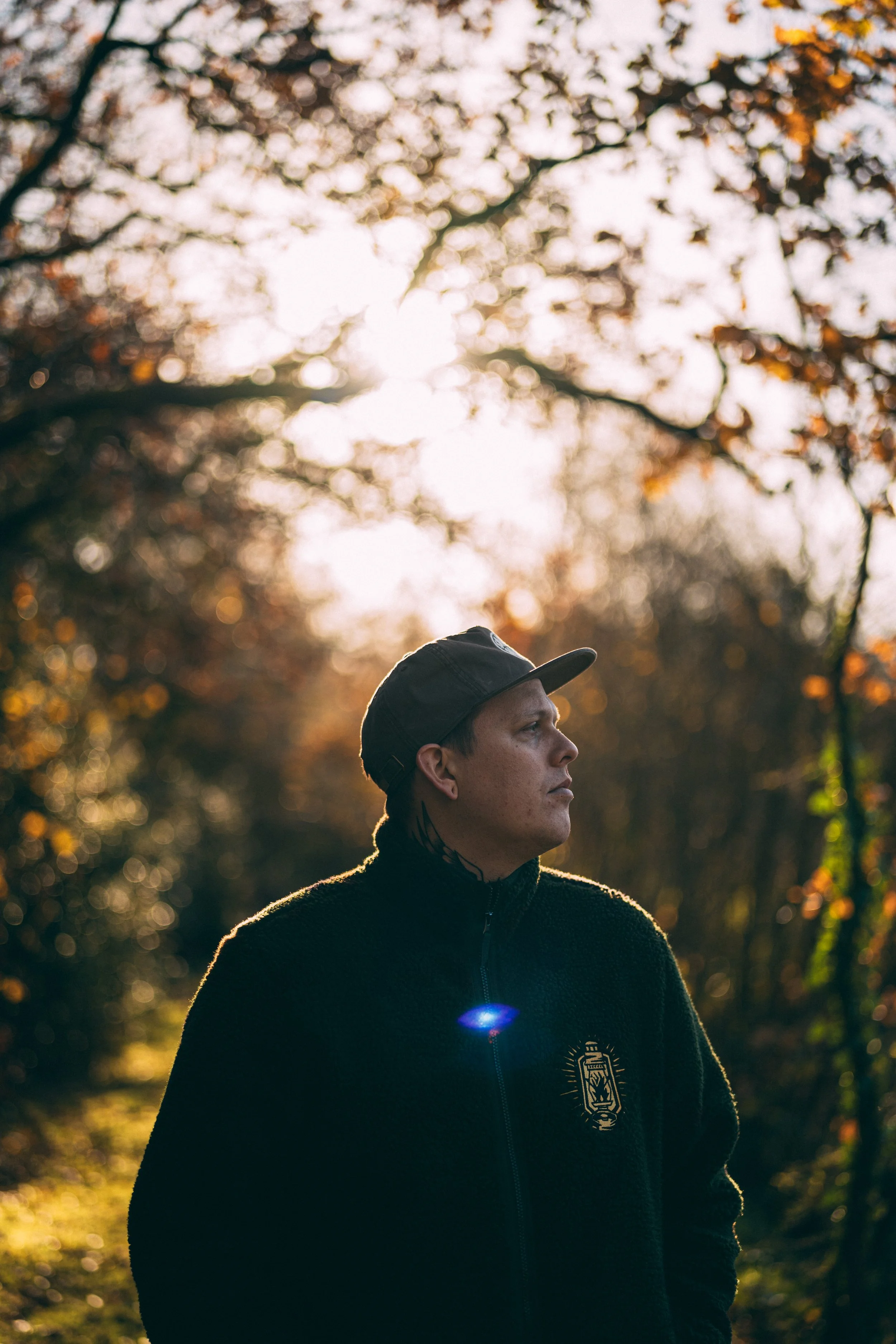 A young man wearing a black jacket and cap standing outdoors in a forest during sunset, with sunlight filtering through autumn leaves.