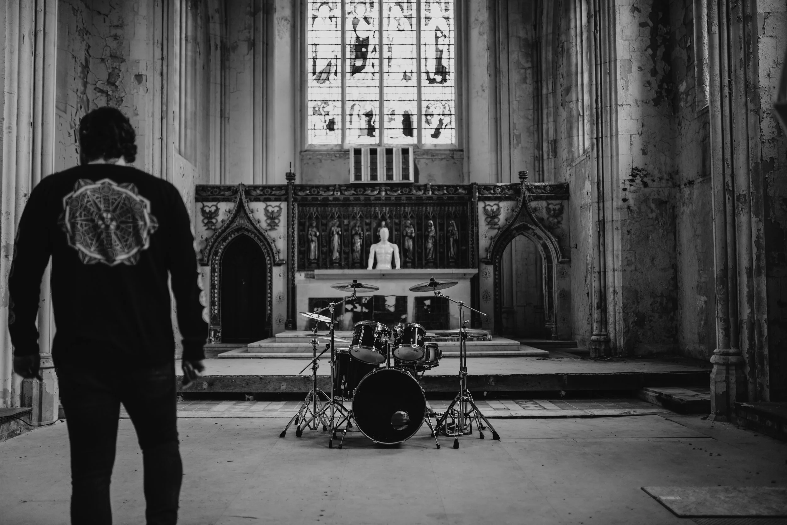 A man walking inside an old, abandoned church with a drum set placed at the front. The church features gothic architecture with stained glass windows and religious statues behind the altar, in black and white.