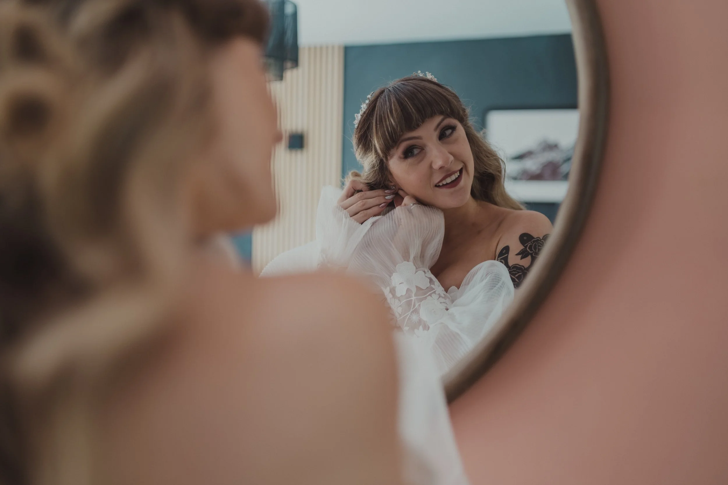 A woman with tattoos on her arm is looking at herself in a mirror, adjusting her earring, with her reflection visible.