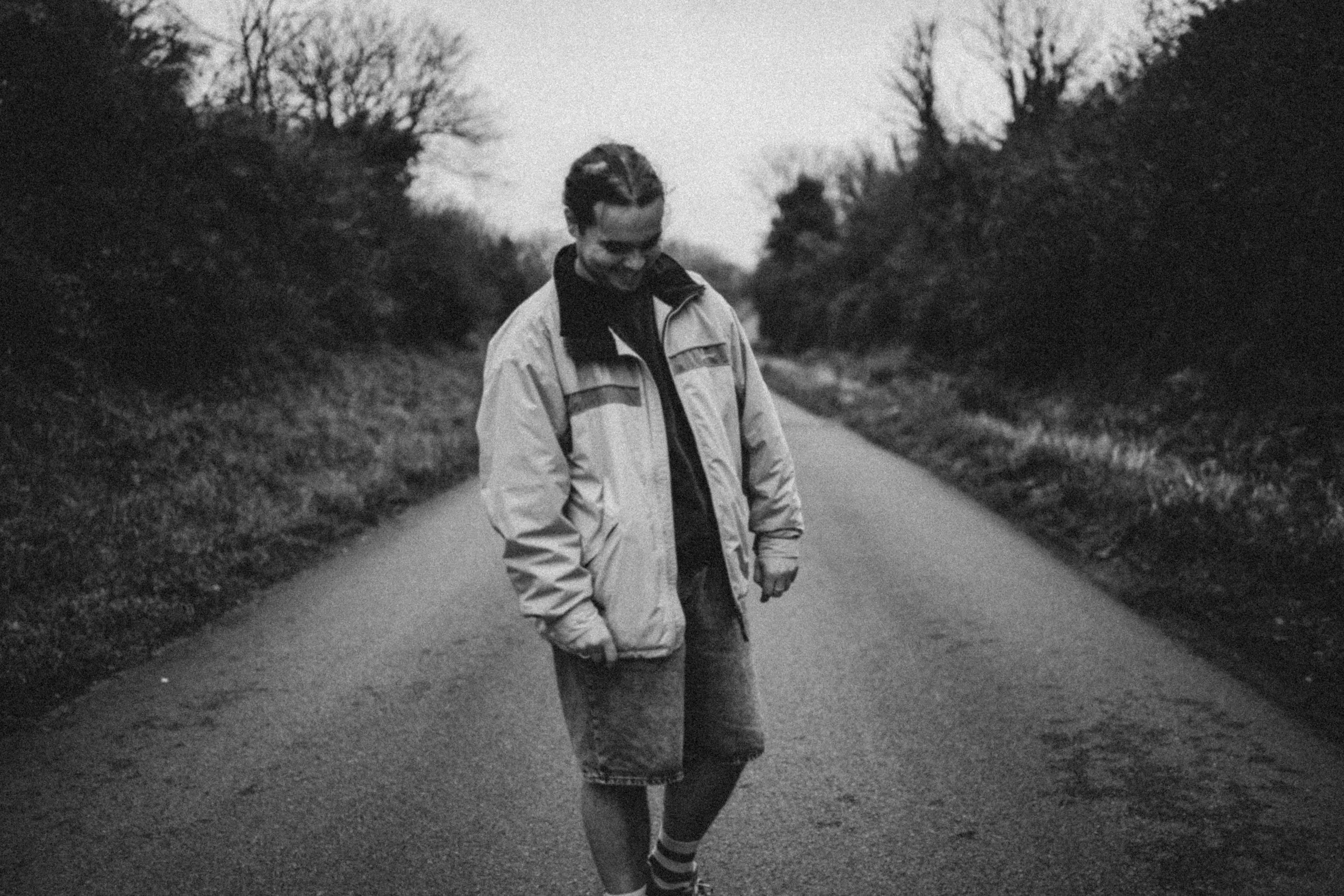 A black-and-white photo of a young man standing on a rural road, looking down and smiling, dressed in casual clothing with a jacket, shorts, and striped socks.