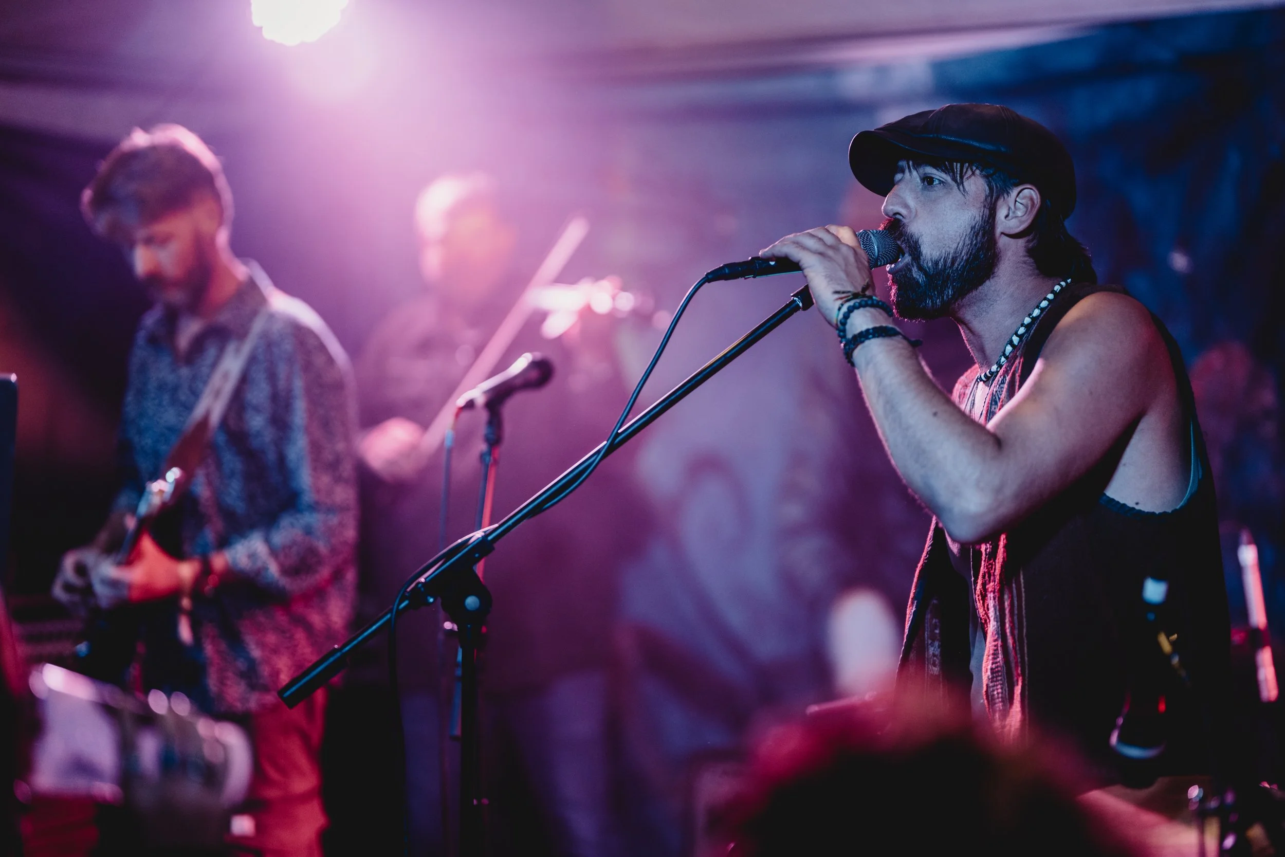 A musician with a beard and wearing a sleeveless top, hat, and jewelry, sings into a microphone on stage with purple and blue lighting, with other band members playing instruments in the background.