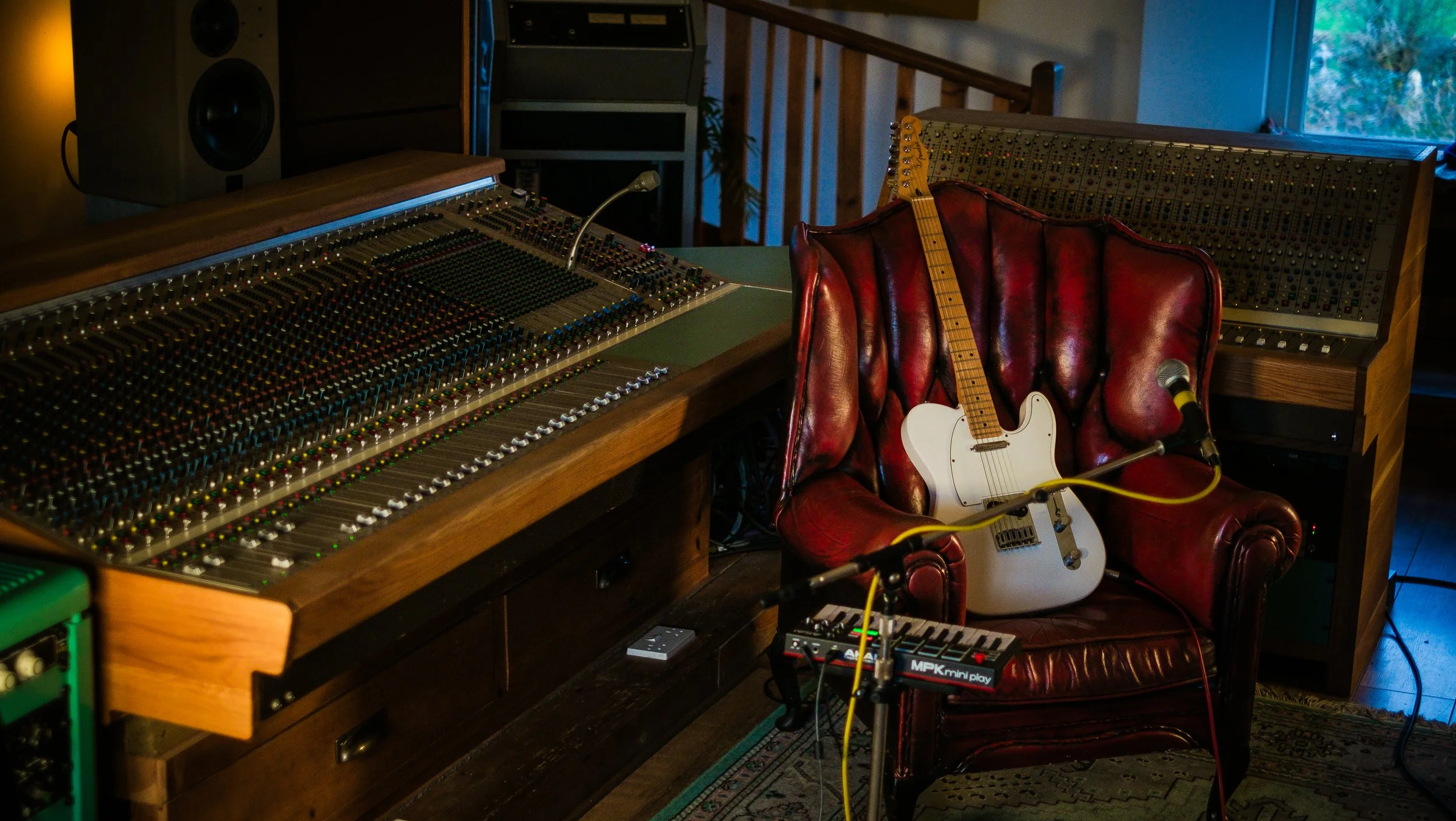Music studio with a large mixing console, an electric guitar on a red leather armchair, and audio equipment, near a window with blue sky visible.
